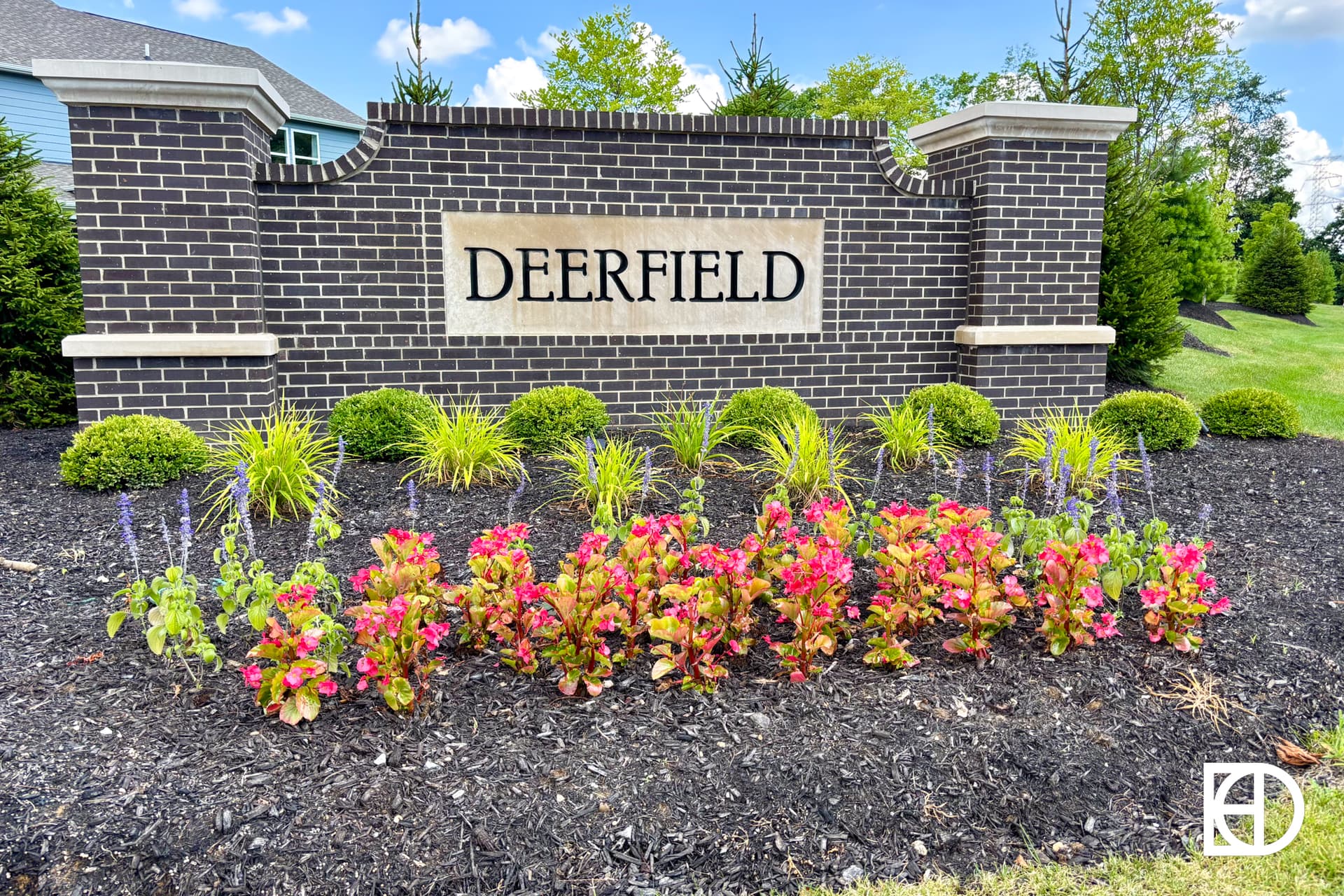 A large brick sign with “DEERFIELD” written on it stands behind neatly landscaped flower beds with colorful flowers and green shrubs, surrounded by grass and trees under a blue sky.