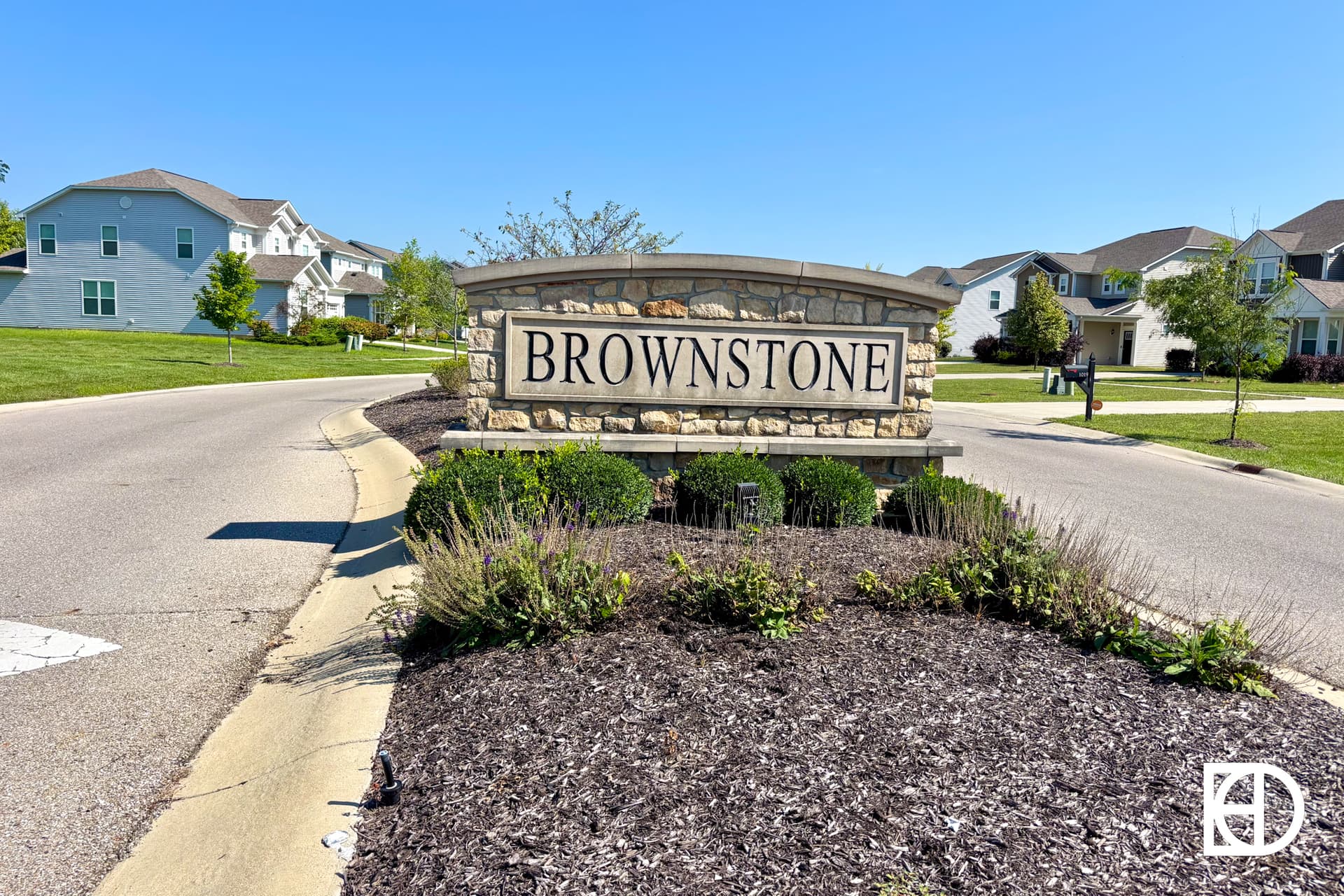 A stone entrance sign with the word BROWNSTONE stands at the entrance of a suburban neighborhood with houses, green lawns, and clear blue sky in the background.