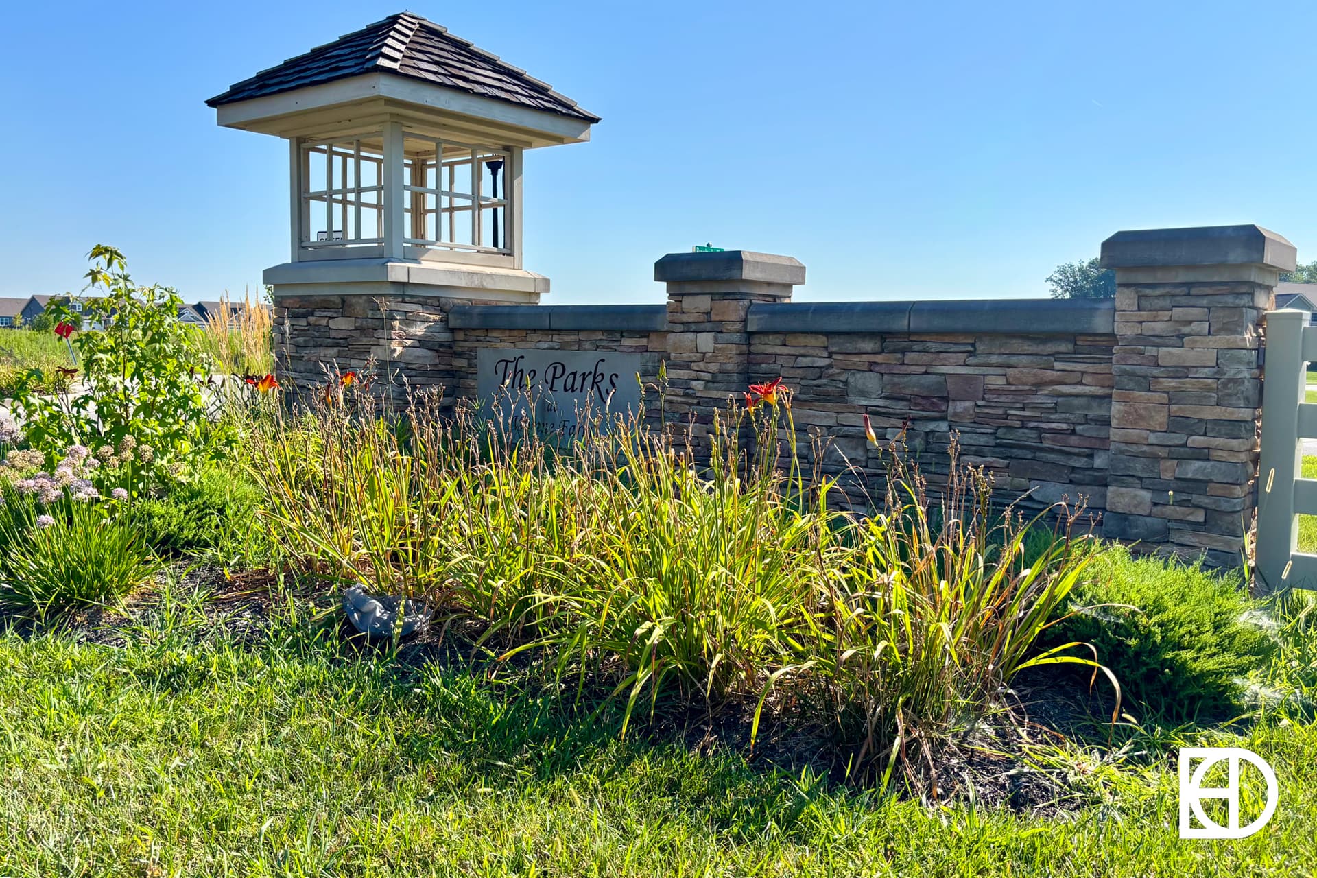 Stone entrance sign with a small roofed structure, surrounded by green grass and blooming plants under a clear blue sky. The sign reads The Parks.