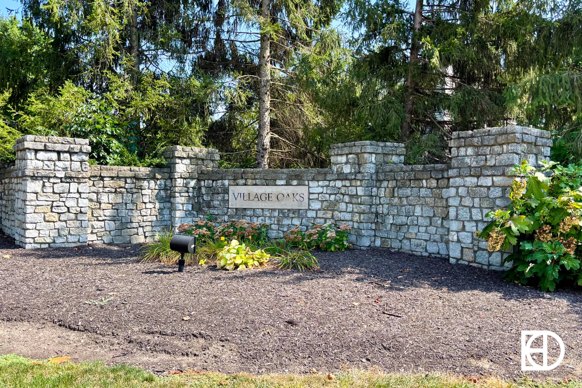 A stone wall with a sign reading VILLAGE OAKS stands among green shrubs and trees, marking the entrance to a neighborhood. A small black mailbox is in front of the wall.