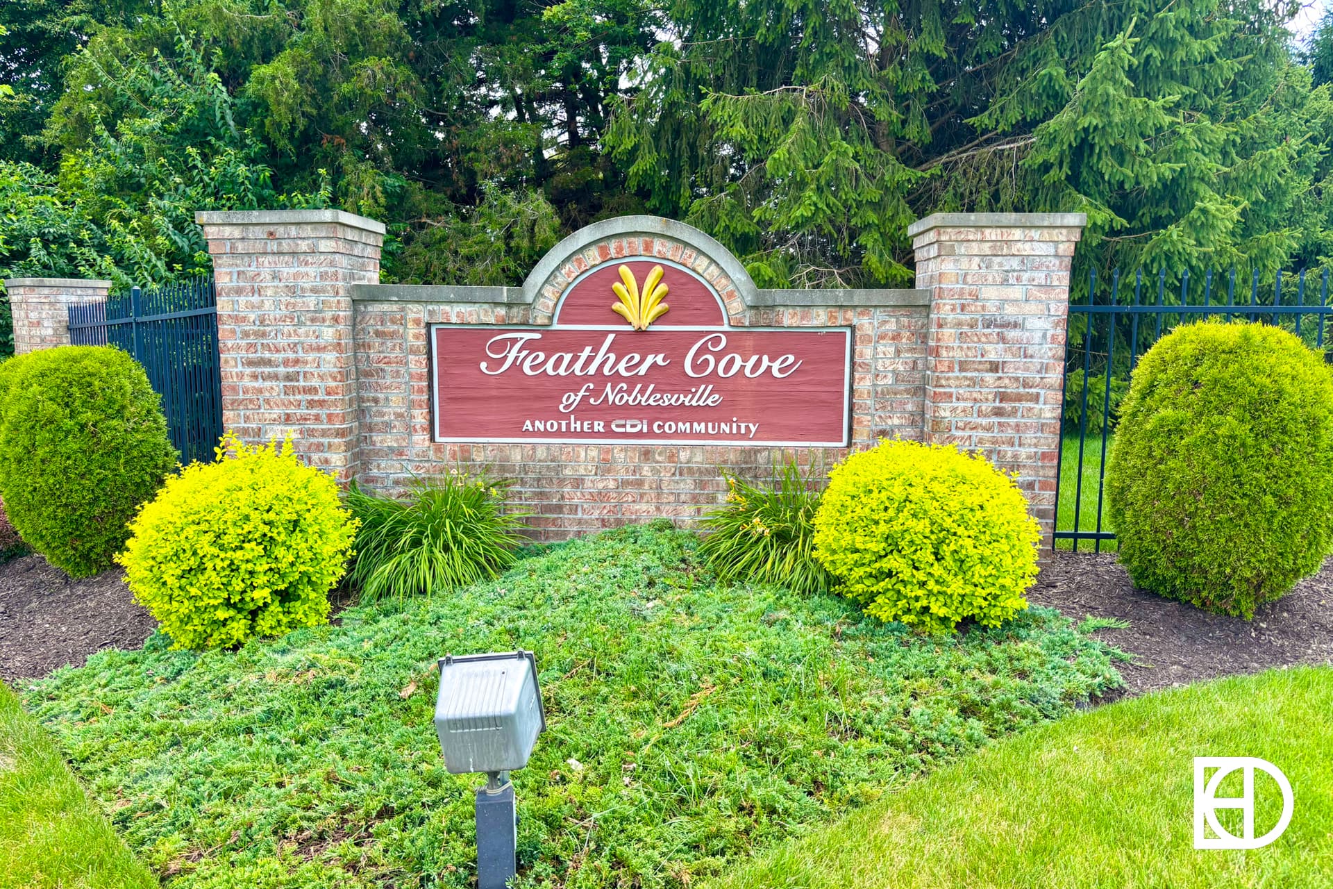 A brick entrance sign reads Feather Cove of Noblesville, Another CDI Community, surrounded by green bushes, grass, and trees with a black fence in the background.