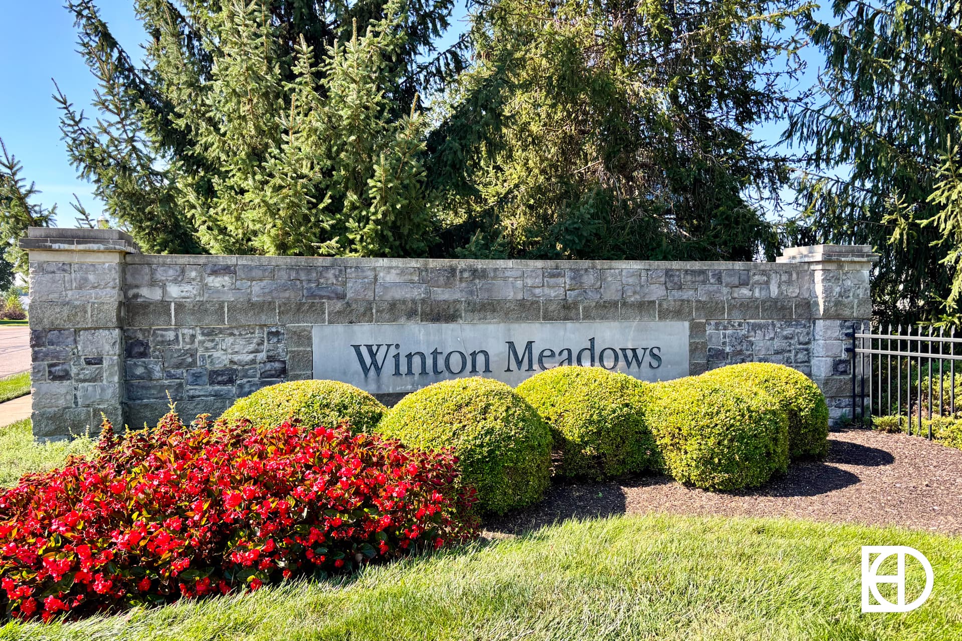 A stone entrance sign reads Winton Meadows, surrounded by manicured bushes, bright red flowers, and tall evergreen trees under a clear blue sky.