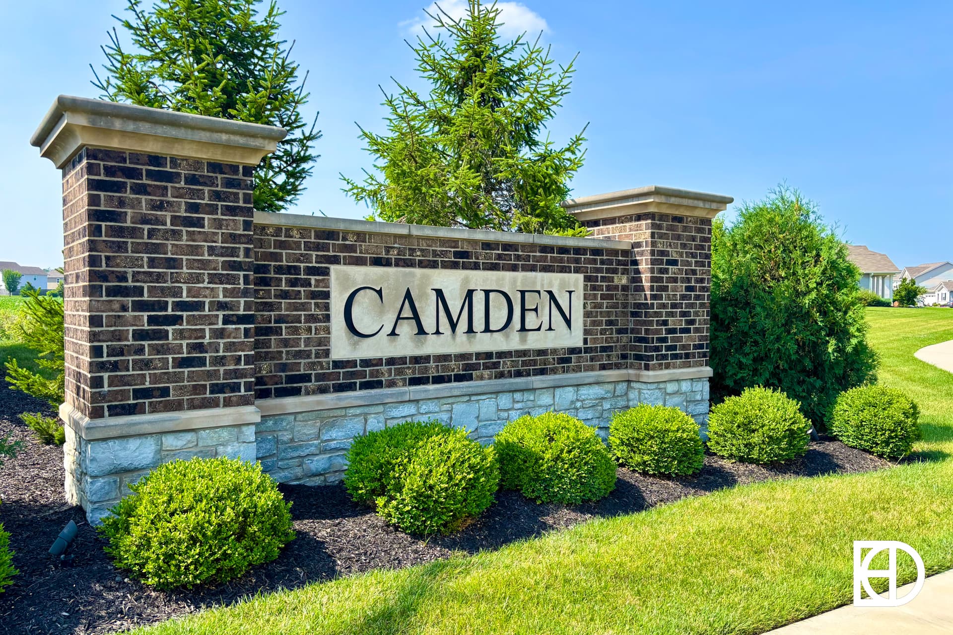 A brick and stone entrance sign reads CAMDEN, surrounded by neatly trimmed bushes, green grass, and trees on a sunny day.