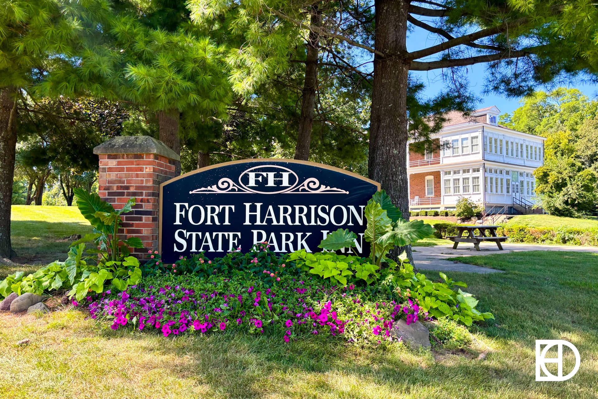 A black sign reading Fort Harrison State Park Inn stands surrounded by flowers and greenery, with a large brick and white building in the background under tall trees.