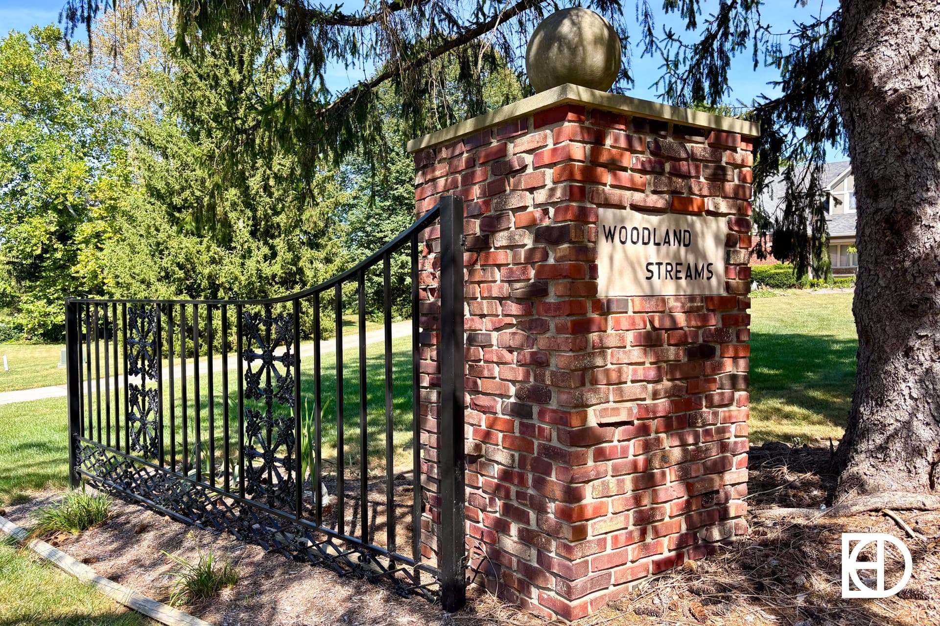 A brick pillar with a stone sphere and a metal gate, featuring a sign that reads “Woodland Streams,” stands on a grassy area surrounded by trees.