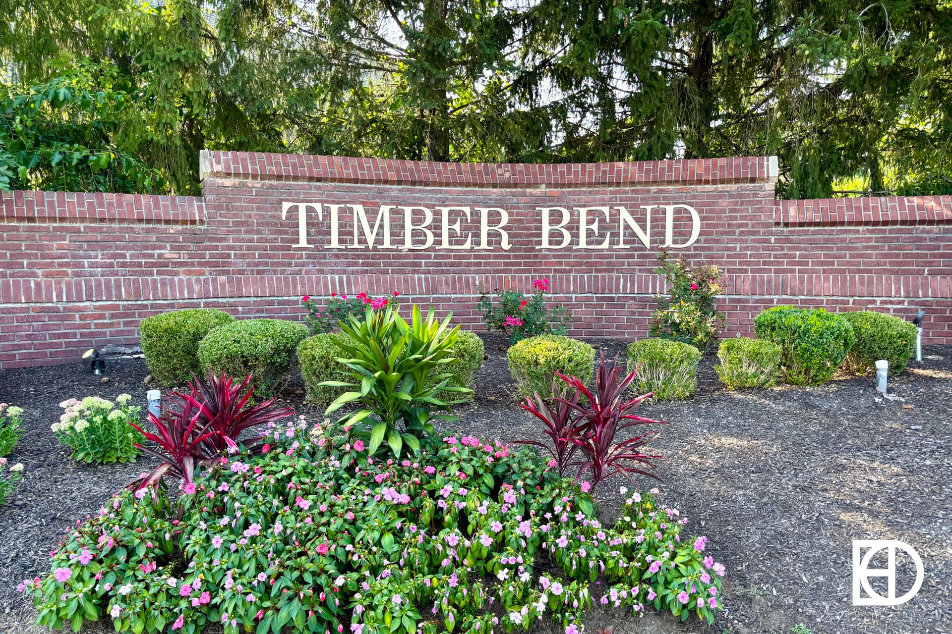 A brick wall with the words TIMBER BEND in large white letters, surrounded by neatly trimmed bushes, colorful flowers, and green plants. Trees and greenery are in the background.