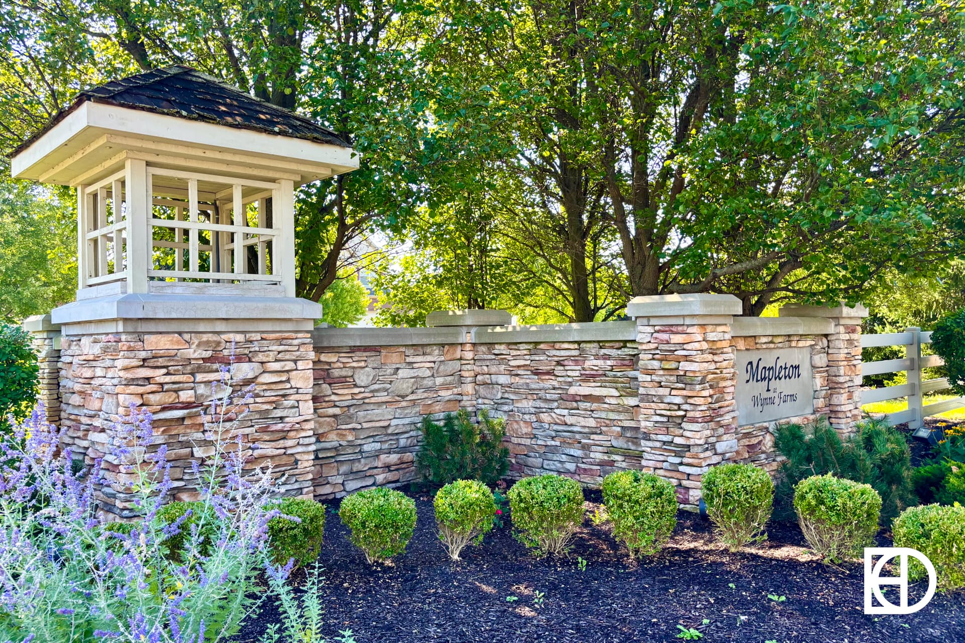 A stone entrance sign for Mapleton Village Series, surrounded by trimmed bushes, purple flowers, and tall green trees, with a gazebo-like structure on top of the stone wall.