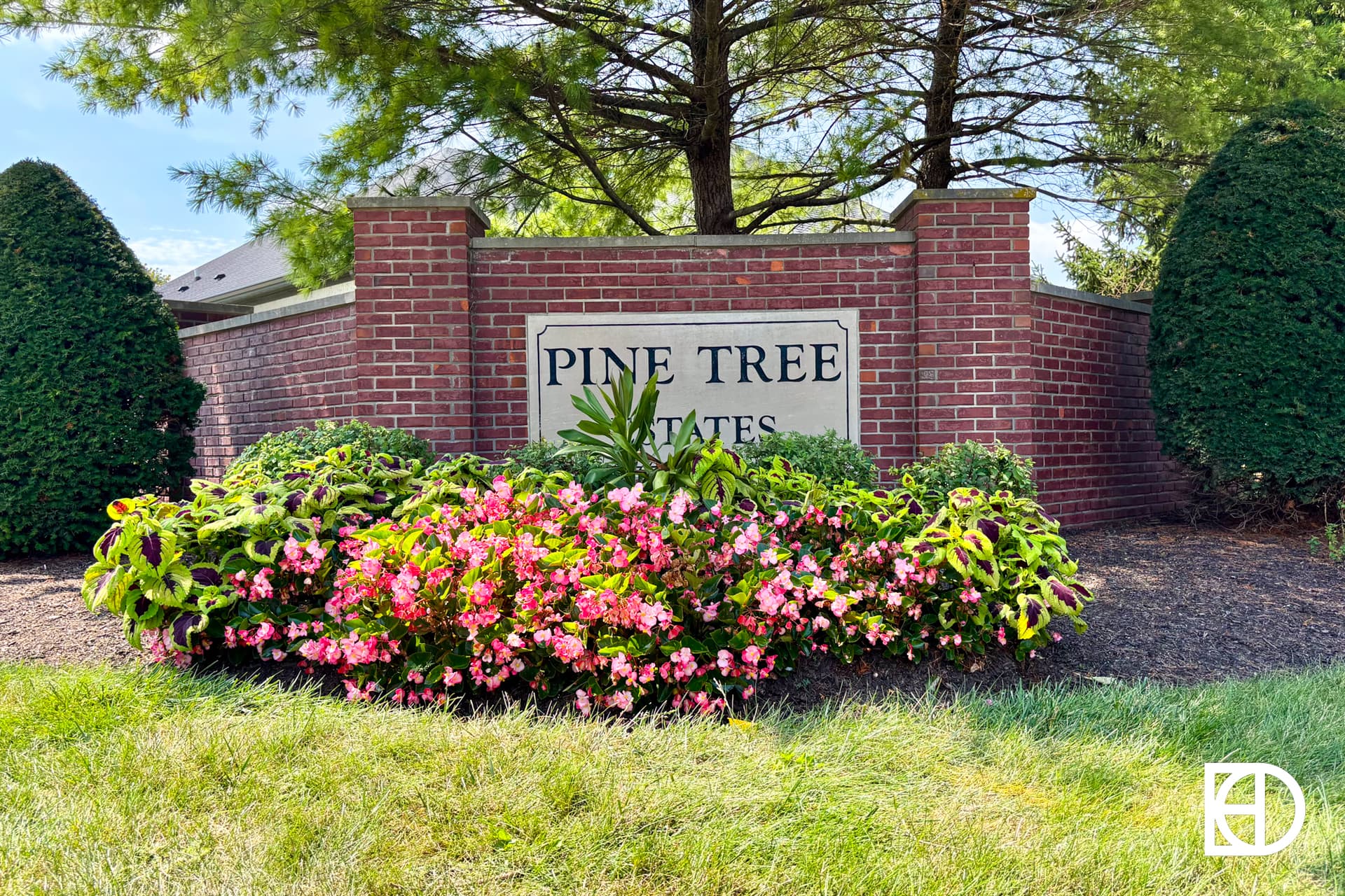 A brick entrance sign for Pine Tree Estates is surrounded by pink flowers, green shrubs, and tall pine trees, with a well-maintained lawn in front.