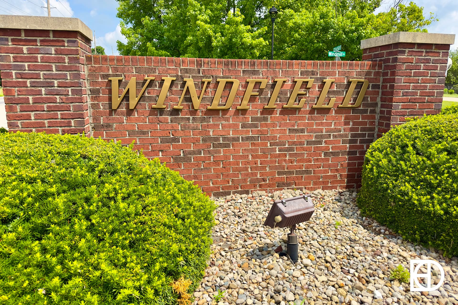 A brick sign with gold letters spelling WINDFIELD stands behind green bushes and landscaping stones, with trees and a blue sky in the background.