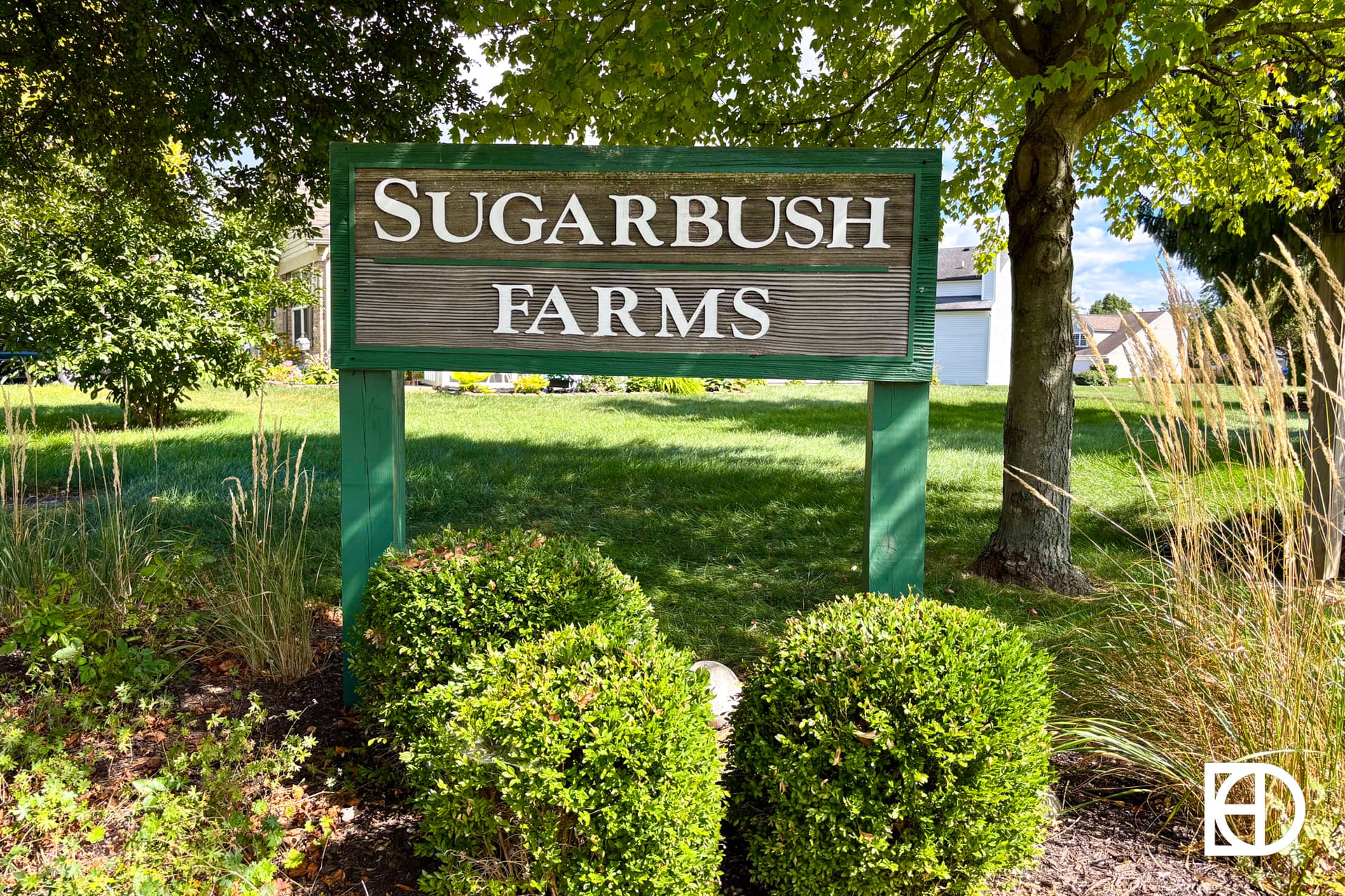 A suburban street with single-story brick houses, well-kept lawns, and trimmed trees under a bright blue sky with scattered white clouds.