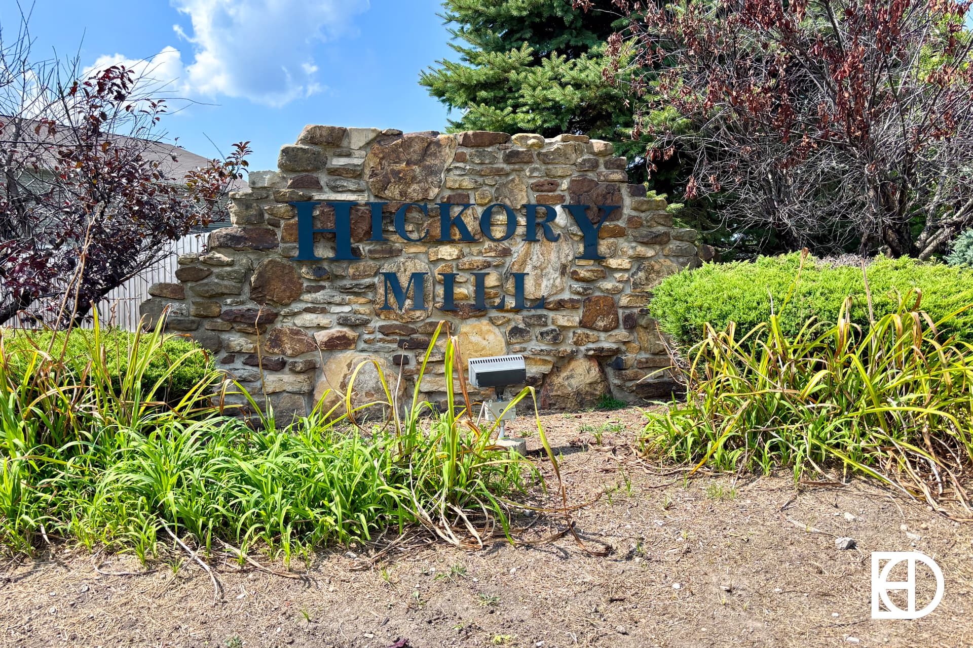 A stone sign with blue letters reading Hickory Mill is surrounded by green shrubs, plants, and trees under a partly cloudy sky.