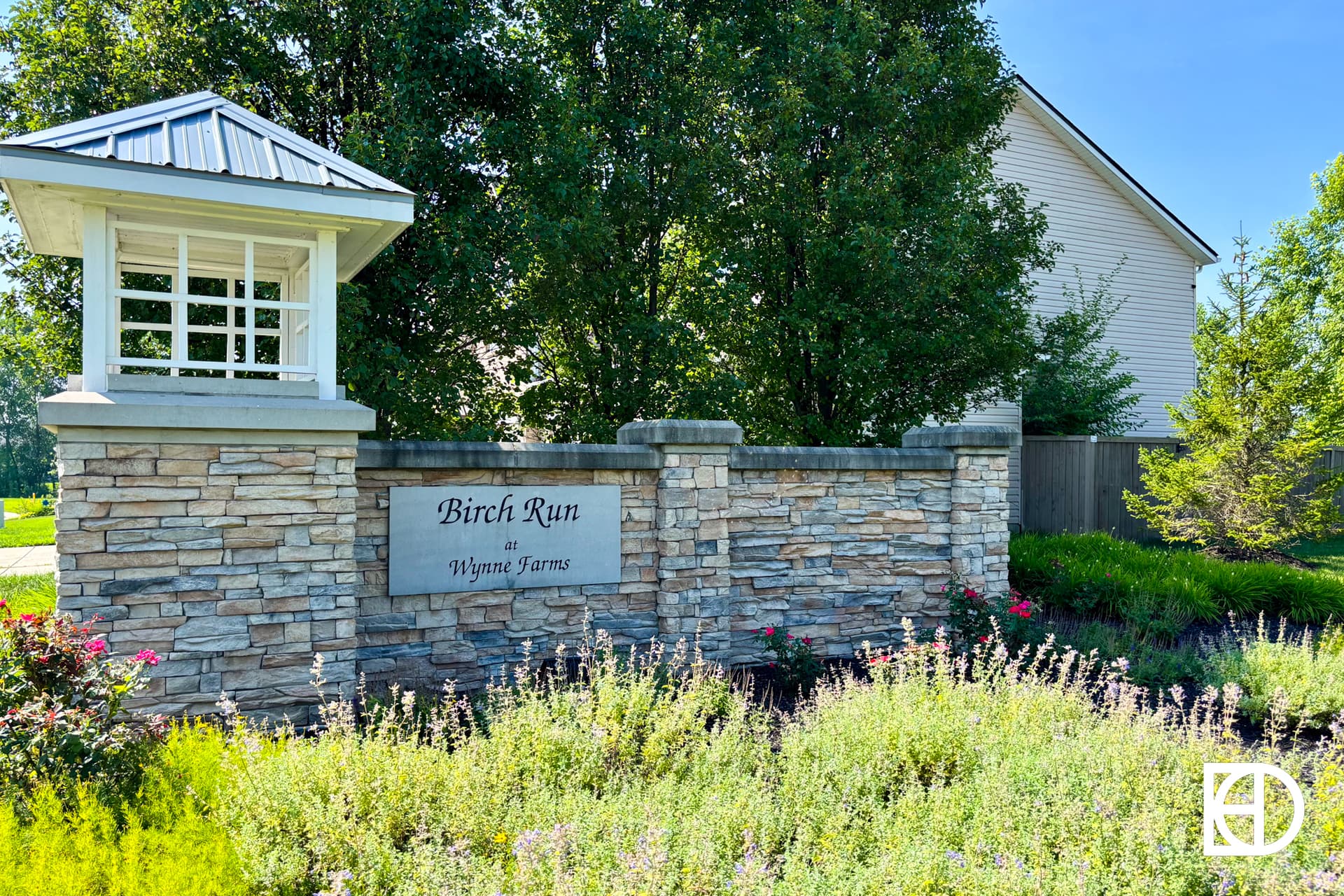 Stone entrance sign for Birch Run at Wynne Farms, surrounded by greenery and flowers, with a small white structure and trees in the background on a sunny day.