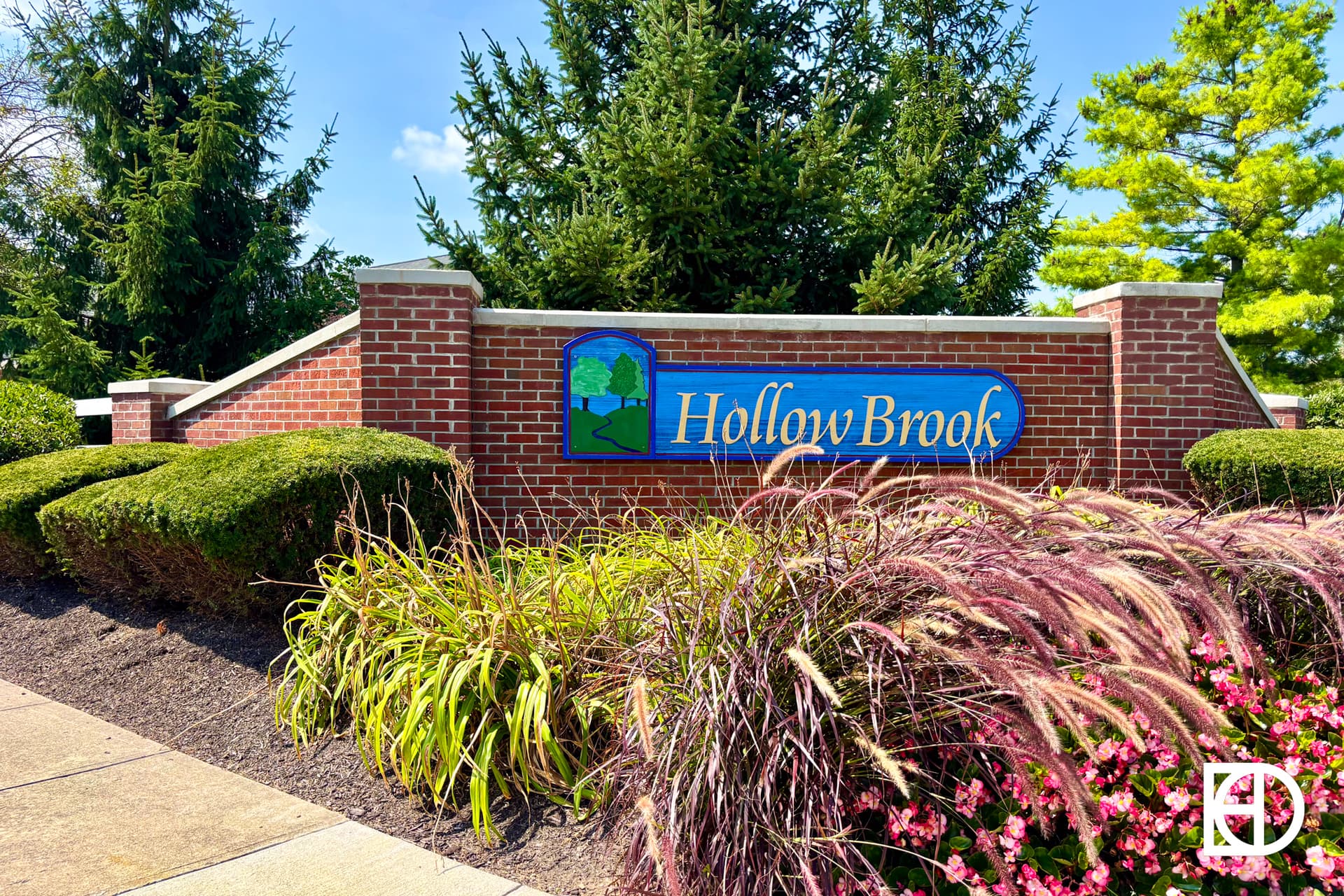 A brick entrance sign for Hollow Brook stands behind manicured bushes, tall grasses, and colorful plants, with evergreen trees and a blue sky in the background.