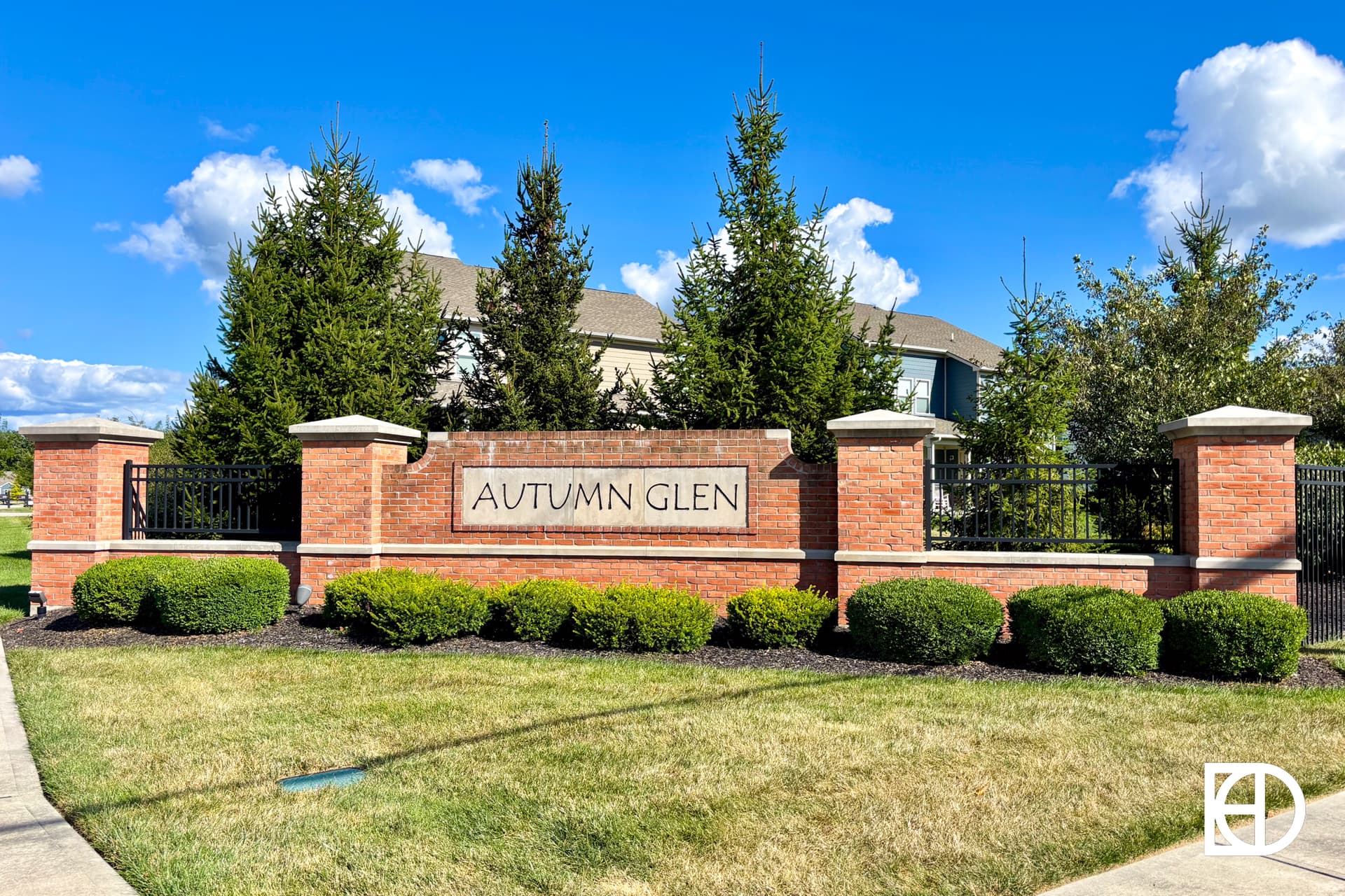 A brick entrance sign with the words AUTUMN GLEN, surrounded by neatly trimmed bushes and trees. A metal fence and residential buildings are visible in the background under a blue sky with clouds