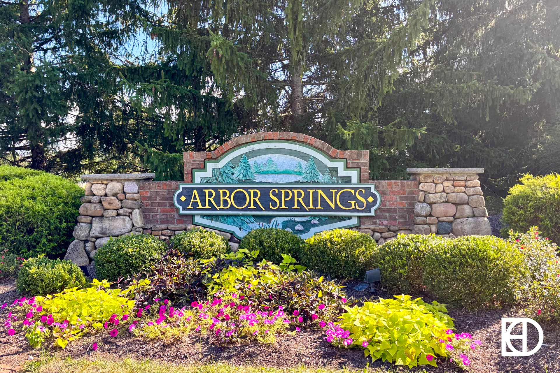 A landscaped stone and brick sign for Arbor Springs surrounded by green bushes, colorful flowers, and tall pine trees in the background.
