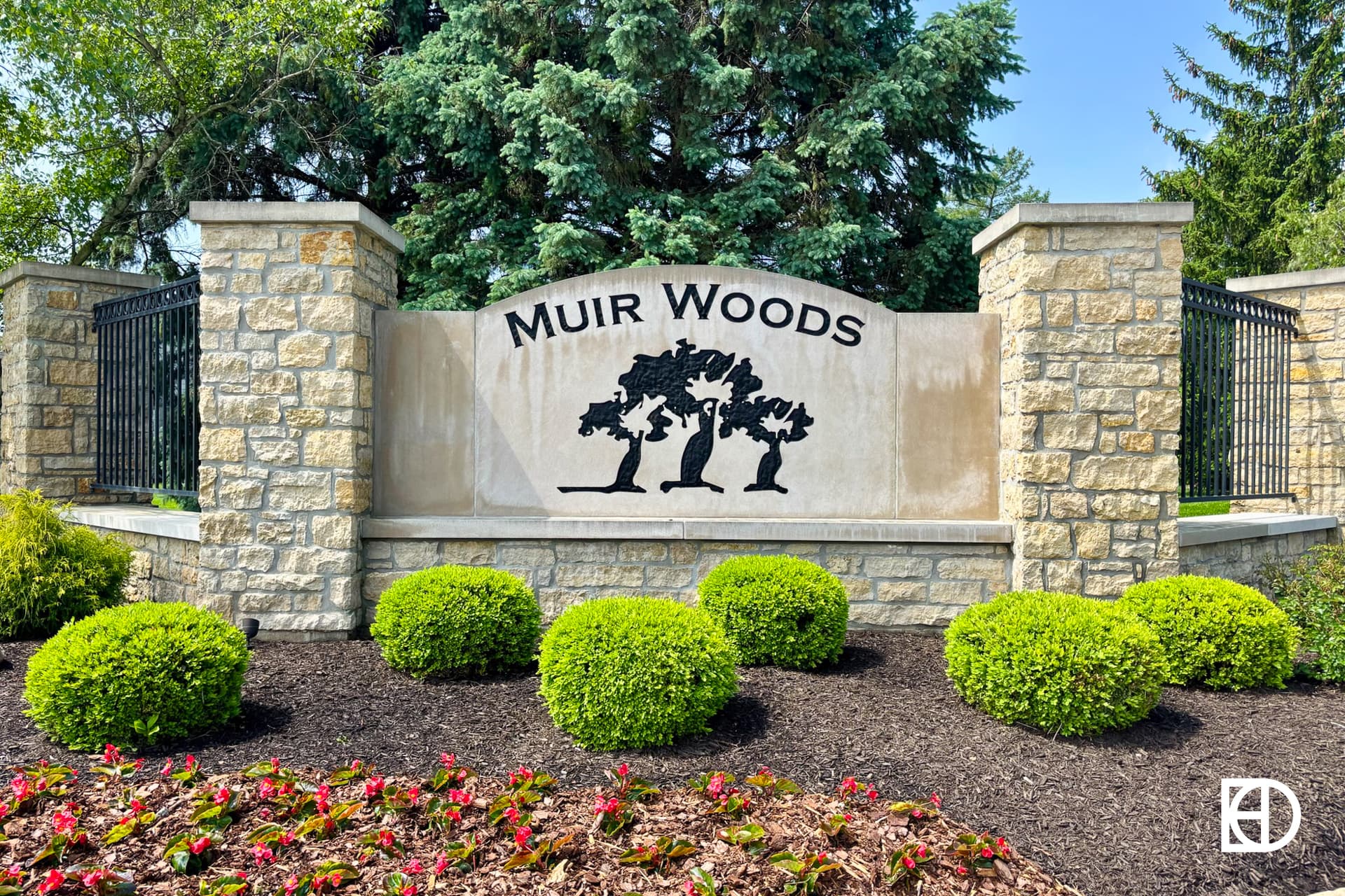 Stone entrance sign for Muir Woods with tree silhouettes, surrounded by green shrubs, flowers, and tall trees in the background.
