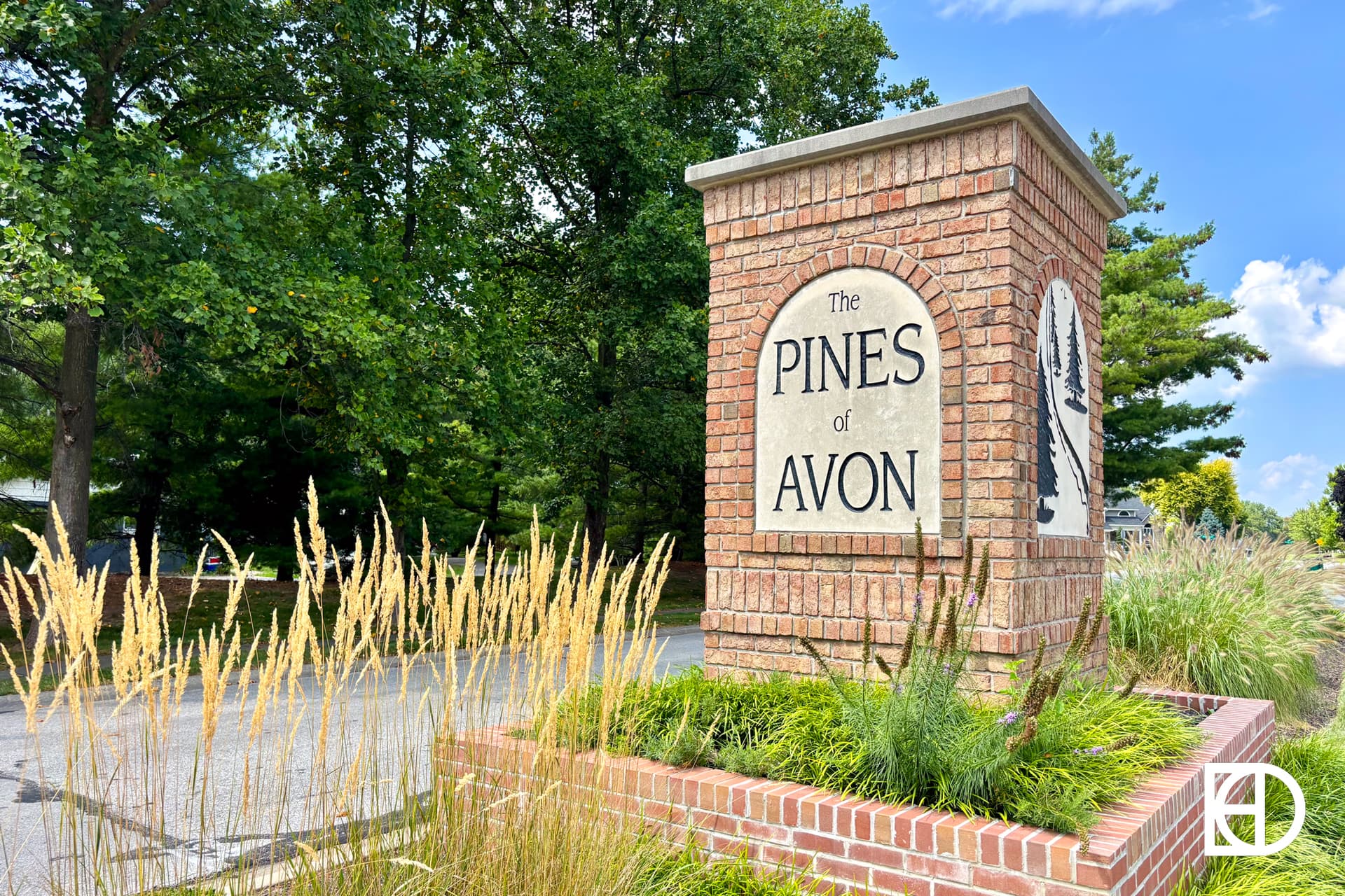A brick entrance sign reads The Pines of Avon surrounded by tall grasses and greenery, with trees and a blue sky in the background.