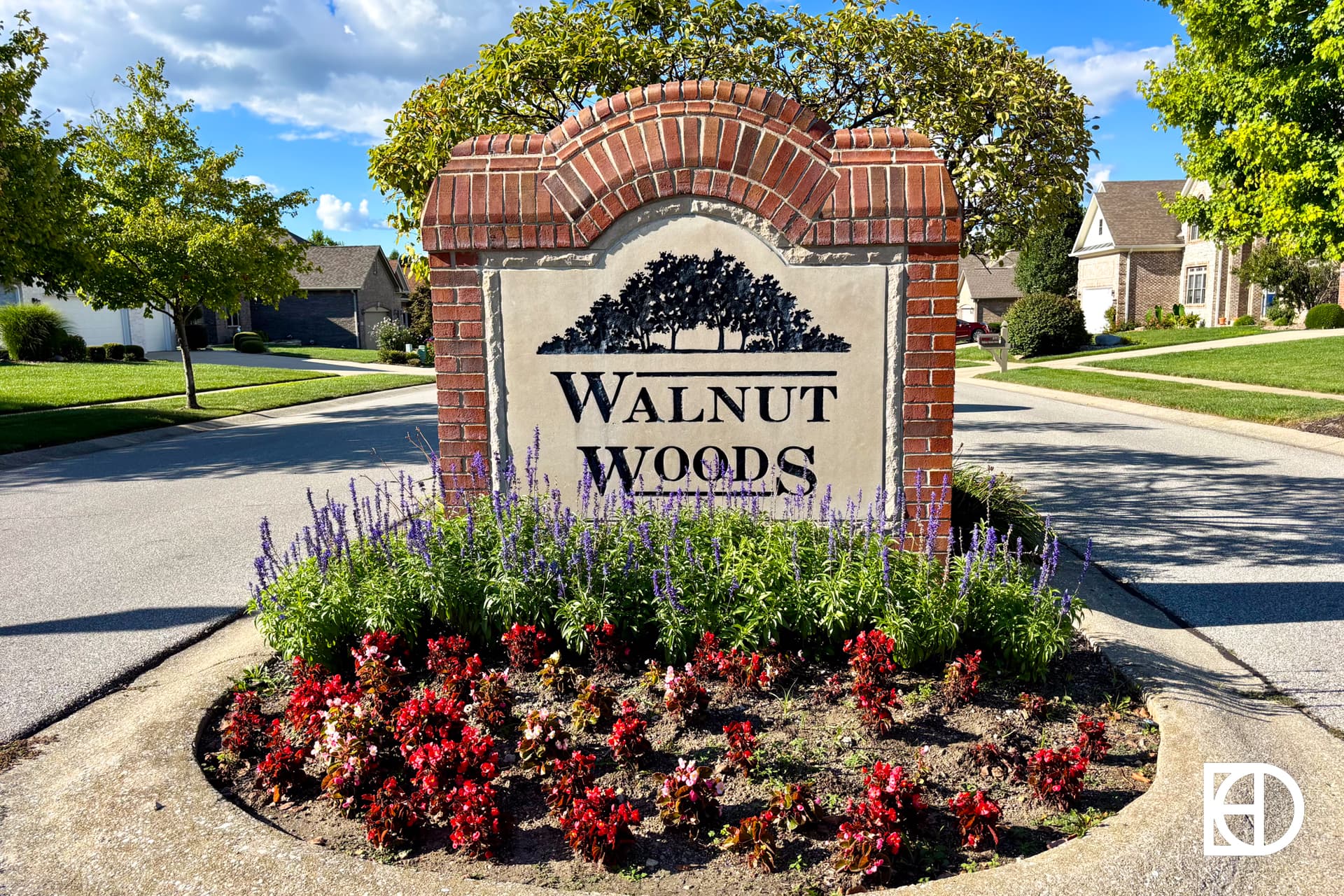 A brick and stone sign reading Walnut Woods stands on a grassy median, surrounded by red and purple flowers, with houses and trees in the background under a blue sky.