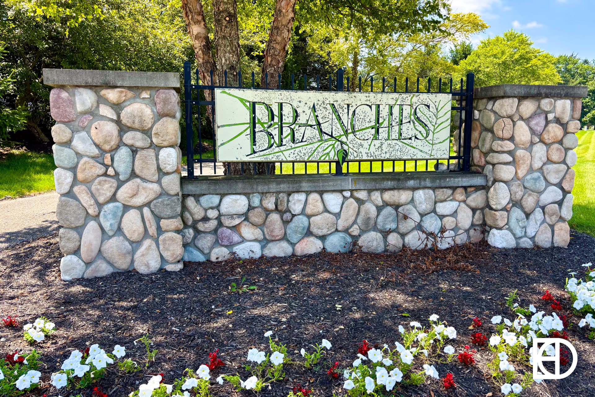 Stone sign with black metal fence reads BRANCHES in green letters with a branch graphic, surrounded by white and red flowers, mulch, and green trees in the background on a sunny day.