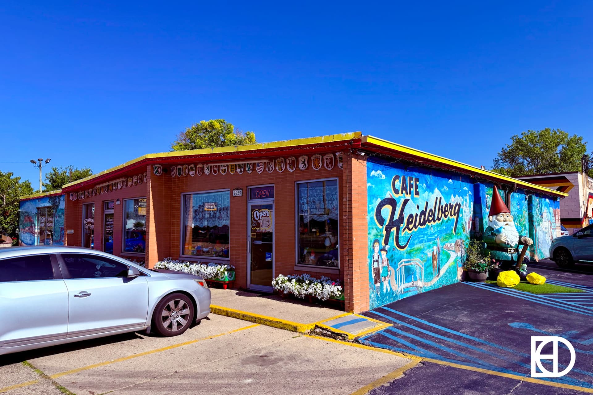 A brick building with a colorful mural reading “Cafe Heidelberg,” featuring a gnome statue outside. A silver car is parked in front, and flower boxes line the windows under a clear blue sky.