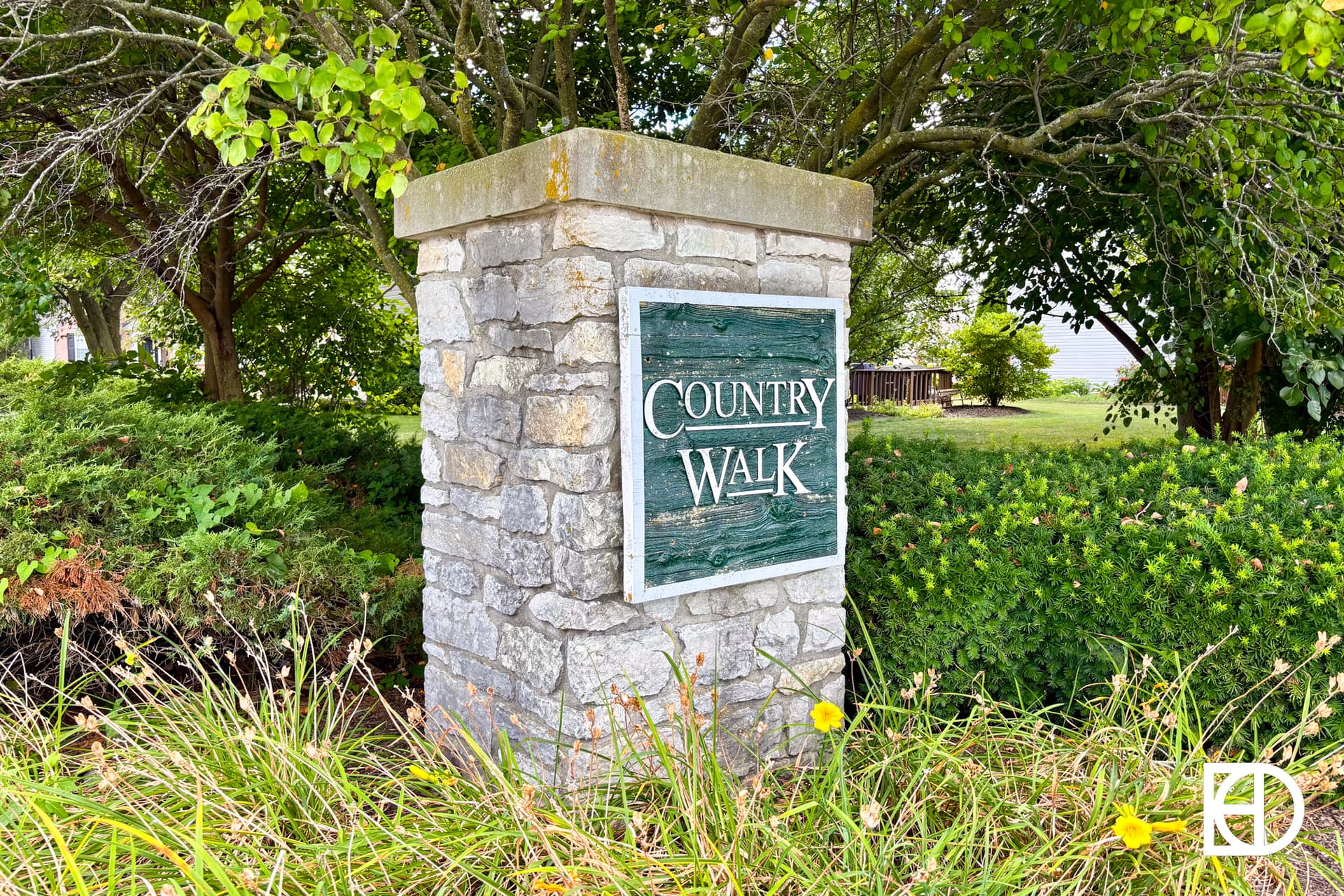 A stone pillar with a weathered green sign reading Country Walk stands among green bushes, grass, and trees. A white logo is visible in the bottom right corner.