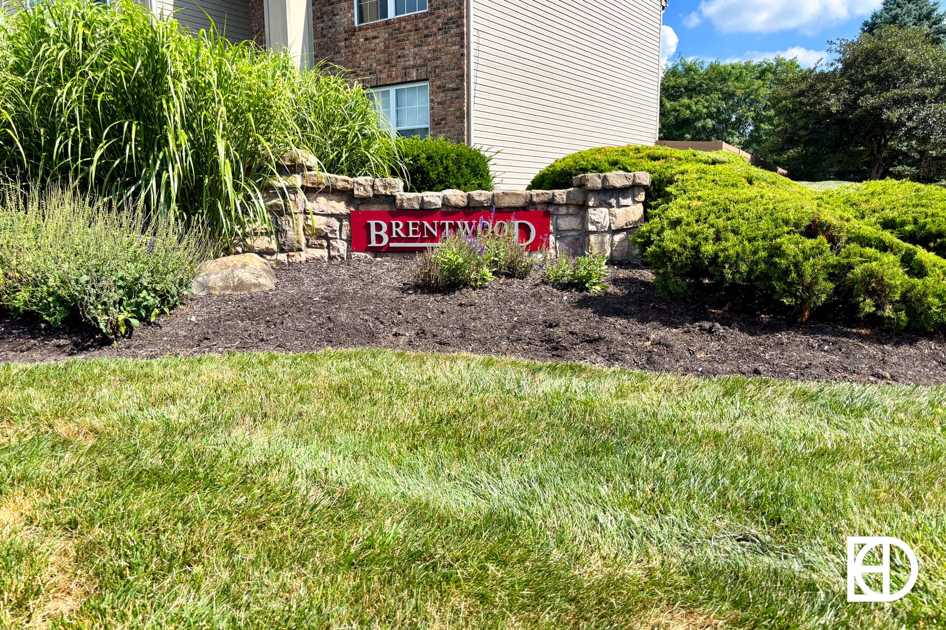 A stone sign reading Brentwood is set in a landscaped garden with bushes and grass in front of a residential building on a sunny day.