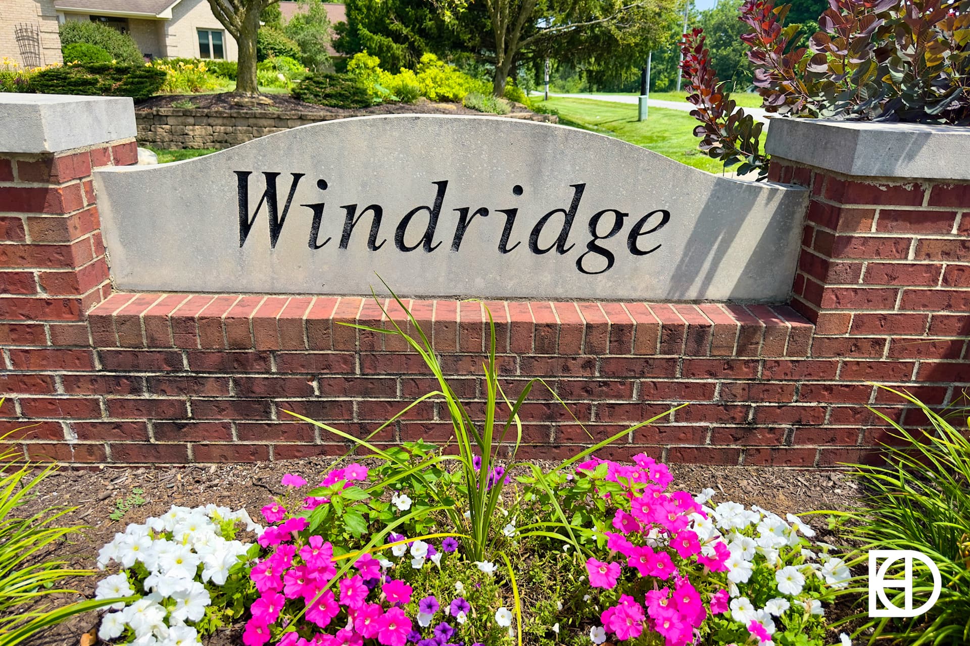 A stone entrance sign reads Birch Run at Wynne Farms, surrounded by green bushes, flowers, and trees, with part of a white building visible in the background on a sunny day.