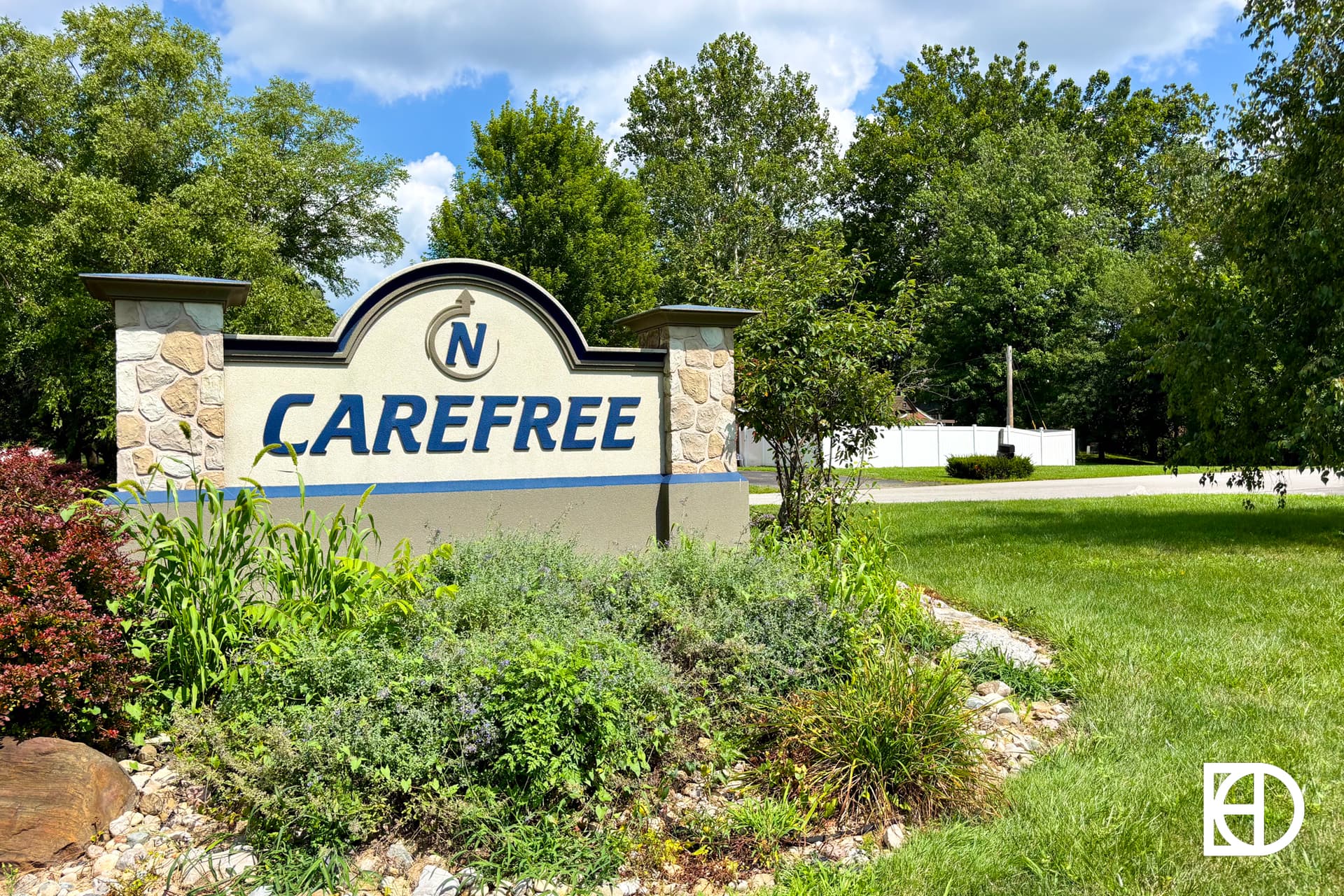 A stone and concrete sign reading CAREFREE stands amid green landscaping, with trees and a sunny sky in the background. A white logo with overlapping letters appears in the bottom right corner.