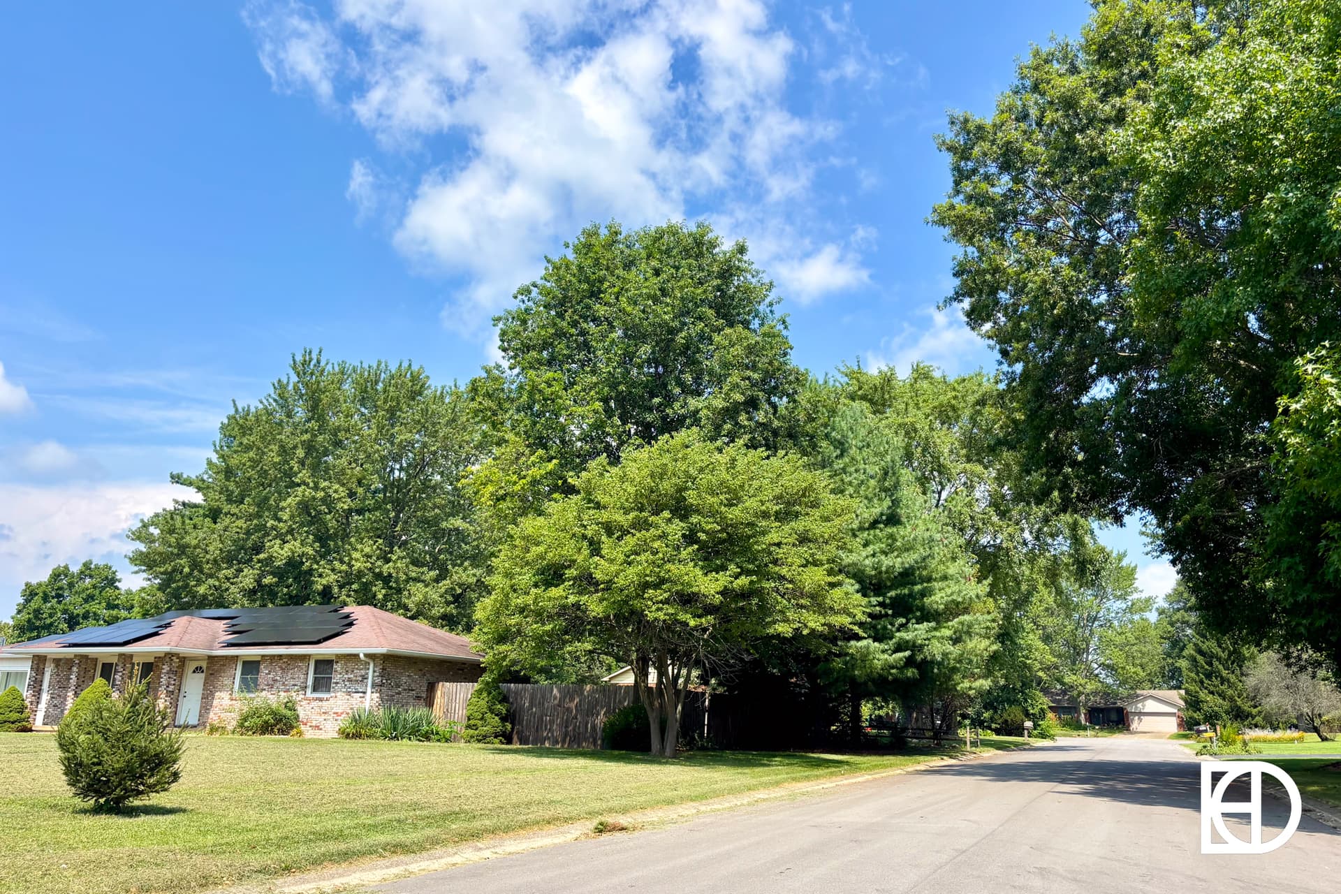 A quiet suburban street with a single-story house on the left, surrounded by lush green trees and lawns under a bright blue sky with scattered clouds. The KD logo appears in the bottom right corner.