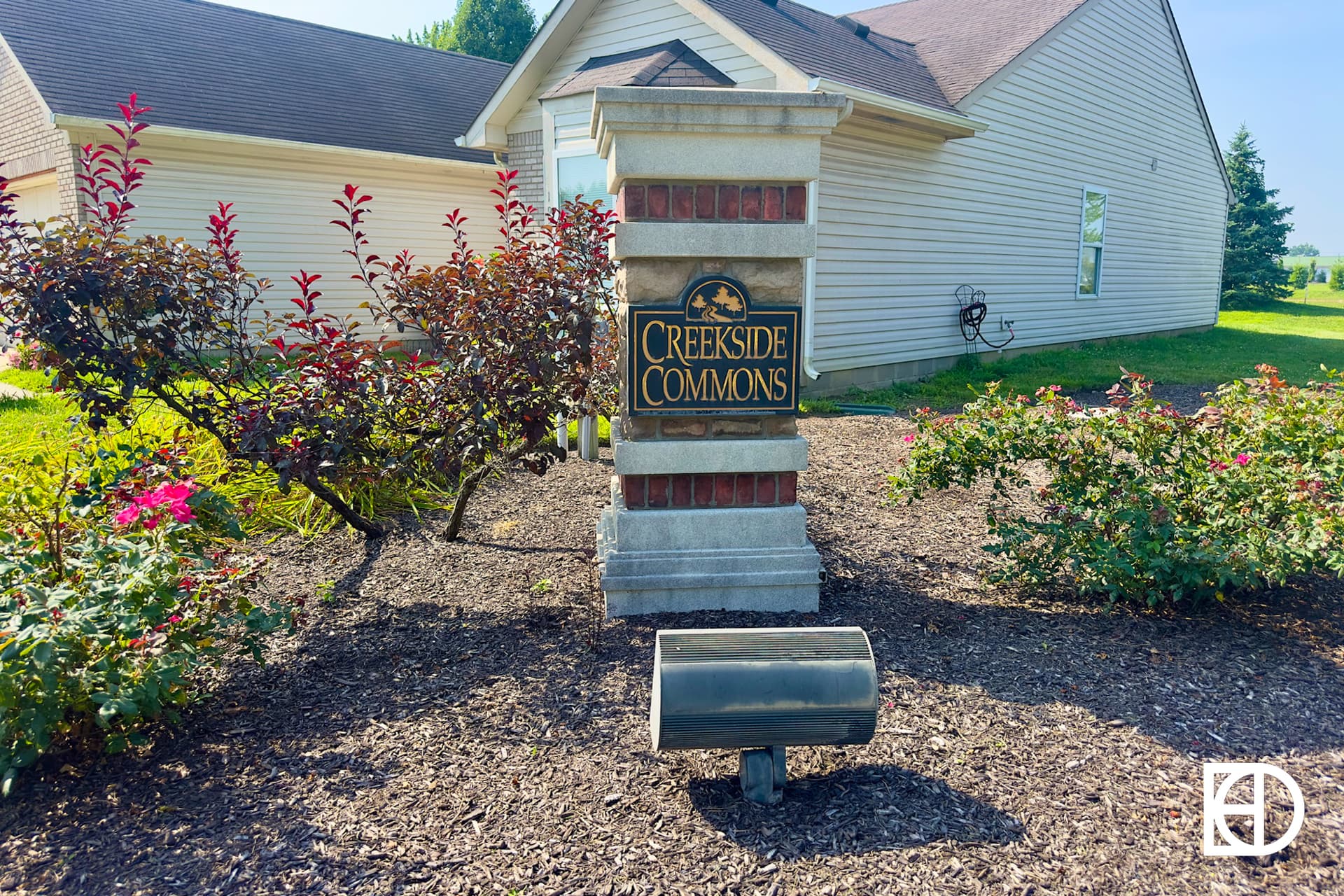 A stone and brick signpost reads “Creekside Commons” in front of landscaped bushes and a beige house with a brown roof. A light fixture is in front of the sign, and the KD logo appears in the bottom right corner.