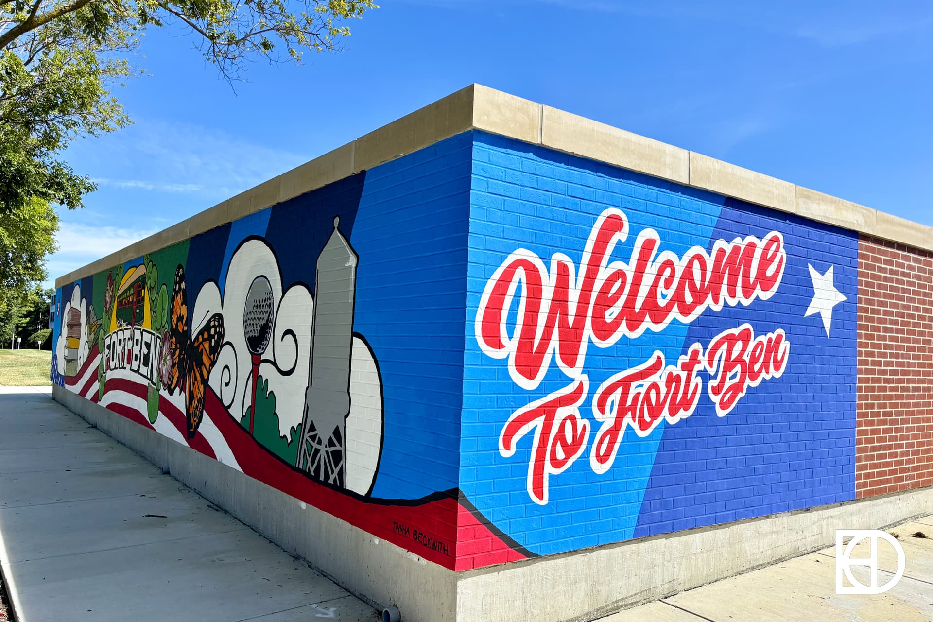 A colorful mural on a brick wall reads Welcome To Fort Ben with images of a water tower, skyline, and local landmarks, set against a blue sky with a white star and red accents.