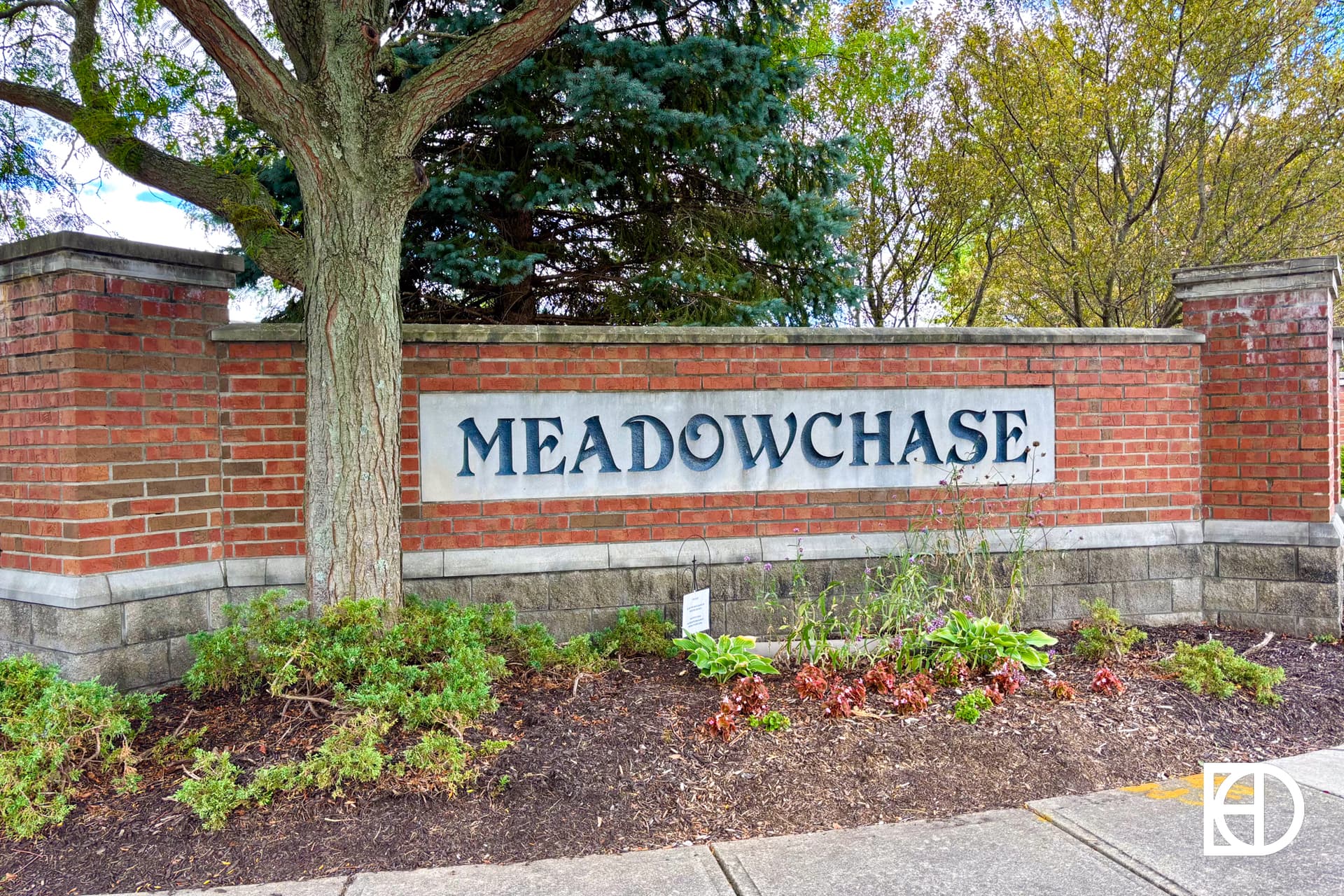 A brick wall with a sign reading MEADOWCHASE in large letters, surrounded by green shrubs, mulch, and trees in the background.