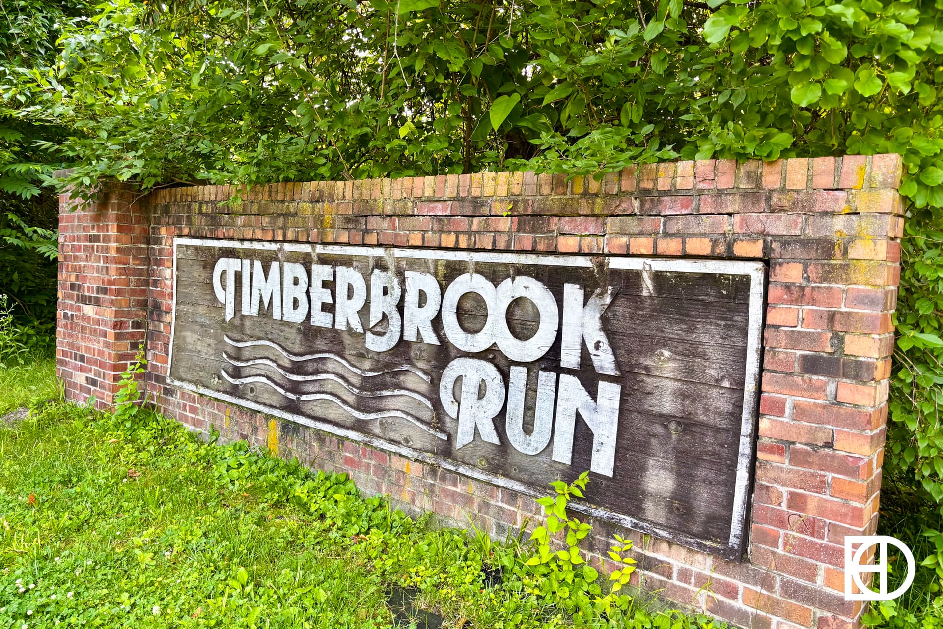 A weathered wooden sign reading Timberbrook Run is mounted on a brick wall surrounded by green grass and lush trees. Wavy lines are painted below the text.