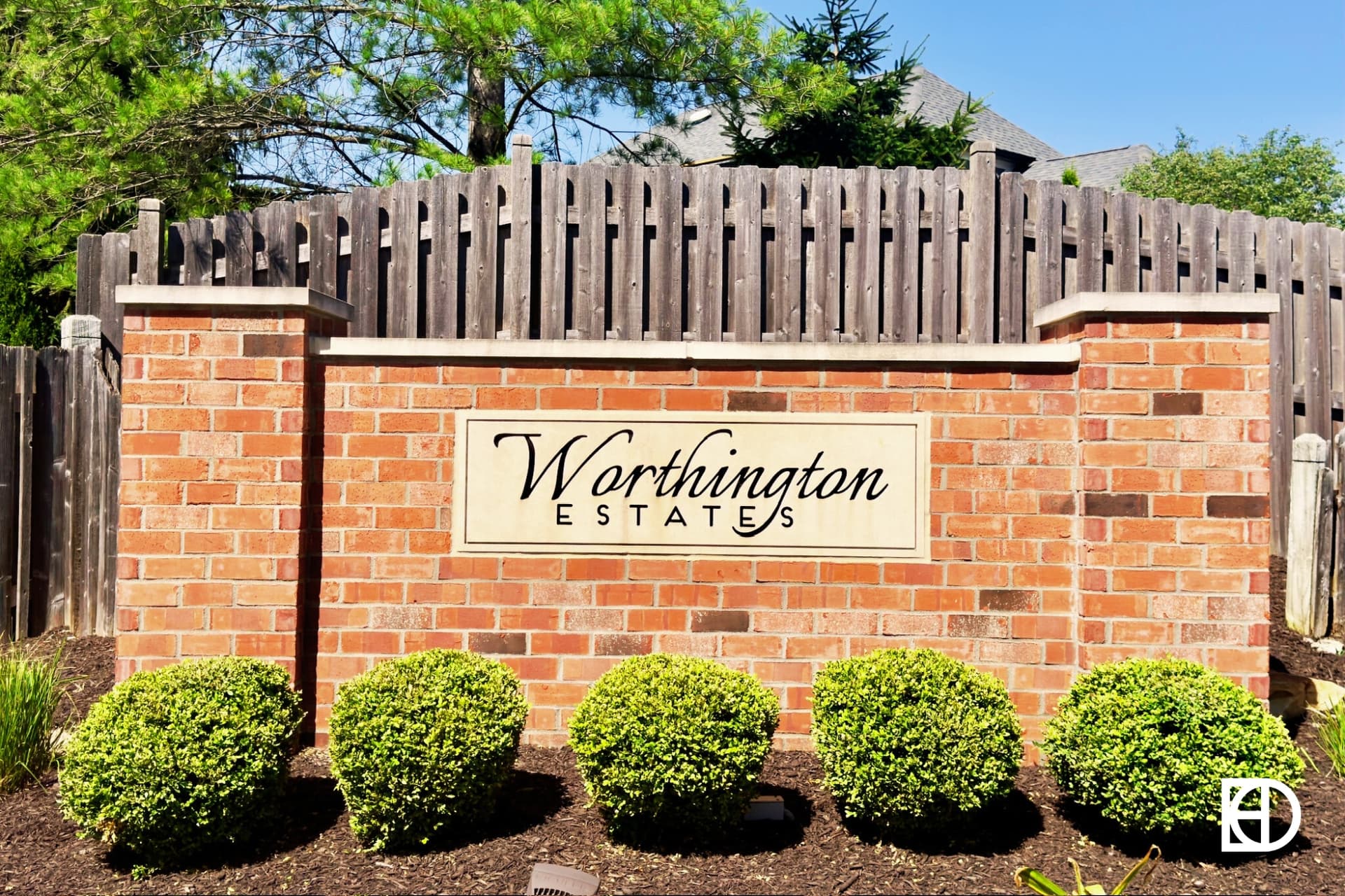 Photo of brick entrance sign for Worthington Estates with small shrubs in front and fence line behind.