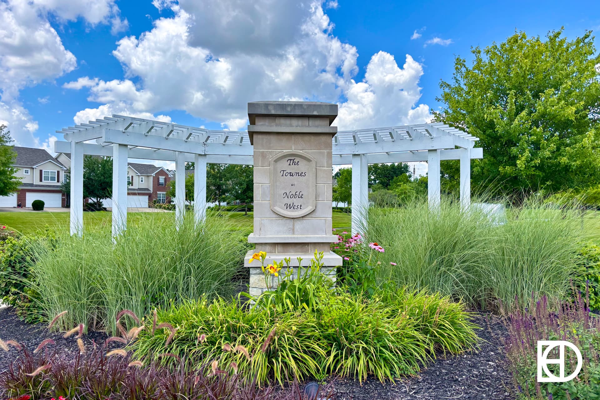 A stone sign reading The Towns at Noble West stands among tall grasses and flowers, in front of a white pergola, with houses and a blue sky with clouds in the background.