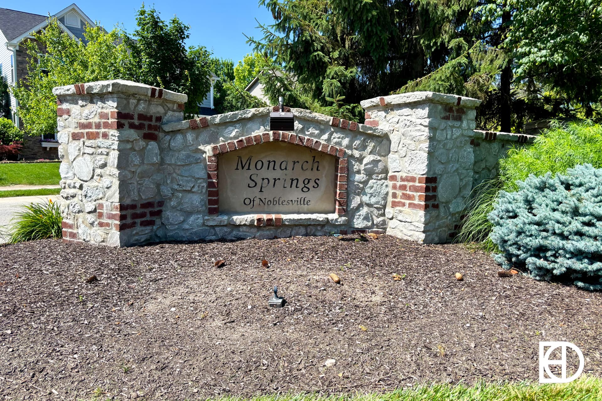 A stone and brick entrance sign reads Monarch Springs of Noblesville, surrounded by mulch, greenery, and trees, with houses visible in the background.