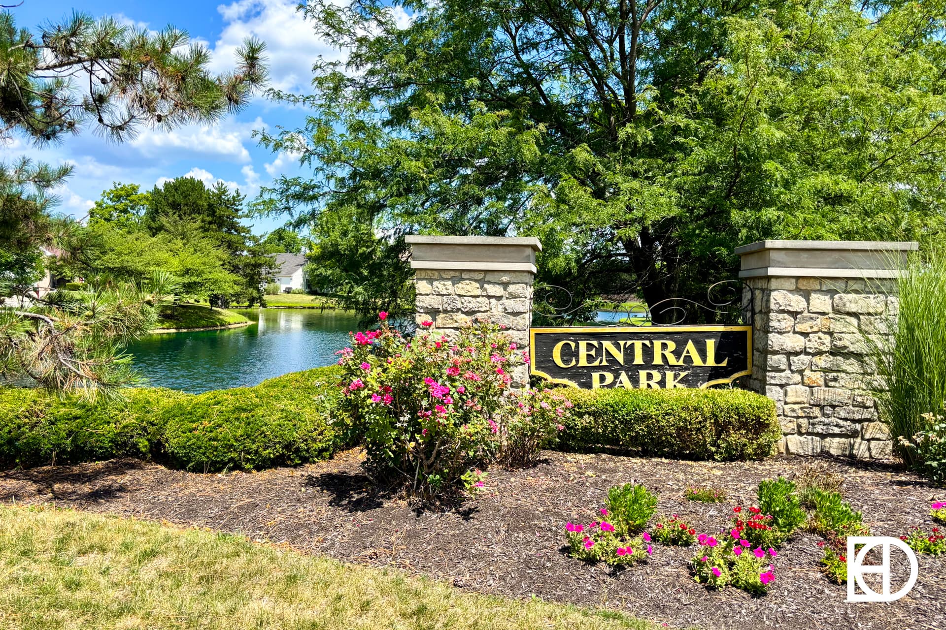 Stone entrance sign reading Central Park surrounded by green bushes, flowering plants, and trees, with a pond and houses visible in the background on a sunny day.