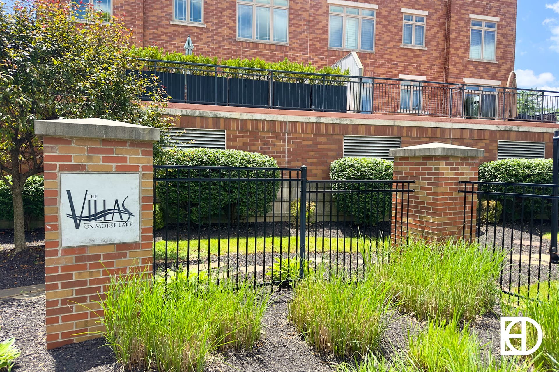 A sunny day at a modern brick apartment complex with balconies, landscaped shrubs, a paved driveway, and parked cars along the side; a blue sky with scattered clouds overhead.
