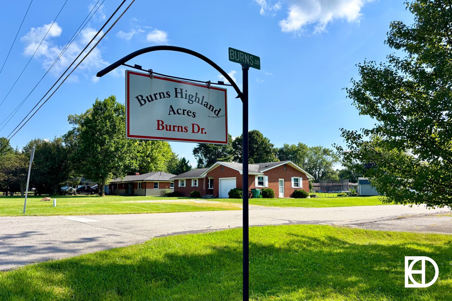 A street sign reads Burns Highland Acres, Burns Dr. near a quiet residential neighborhood with single-story brick houses, green lawns, trees, and a clear blue sky in the background.