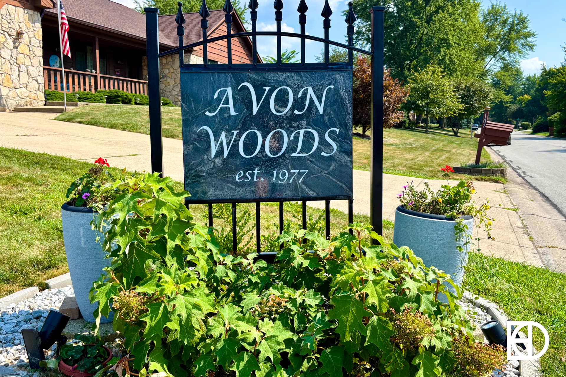 A black metal sign reading “Avon Woods est. 1977” stands among green plants and flowers, near a suburban street with houses, trees, and an American flag in the background.