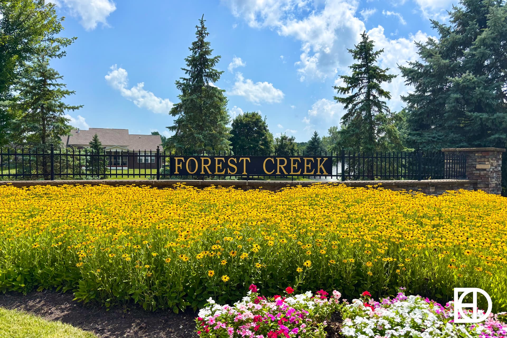 A large bed of yellow flowers and some pink and white flowers in front of a black sign with gold letters reading FOREST CREEK, with pine trees, a black fence, and houses in the background under a blue sky with clouds.