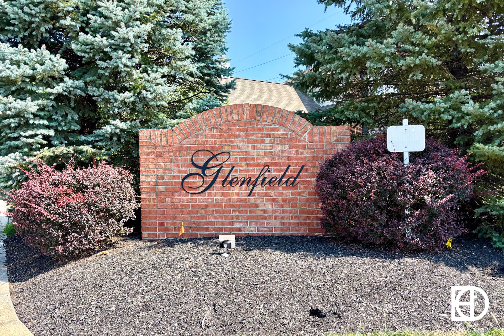 A brick sign with the word Glenfield in cursive, surrounded by bushes and pine trees, stands in a landscaped area with mulch.