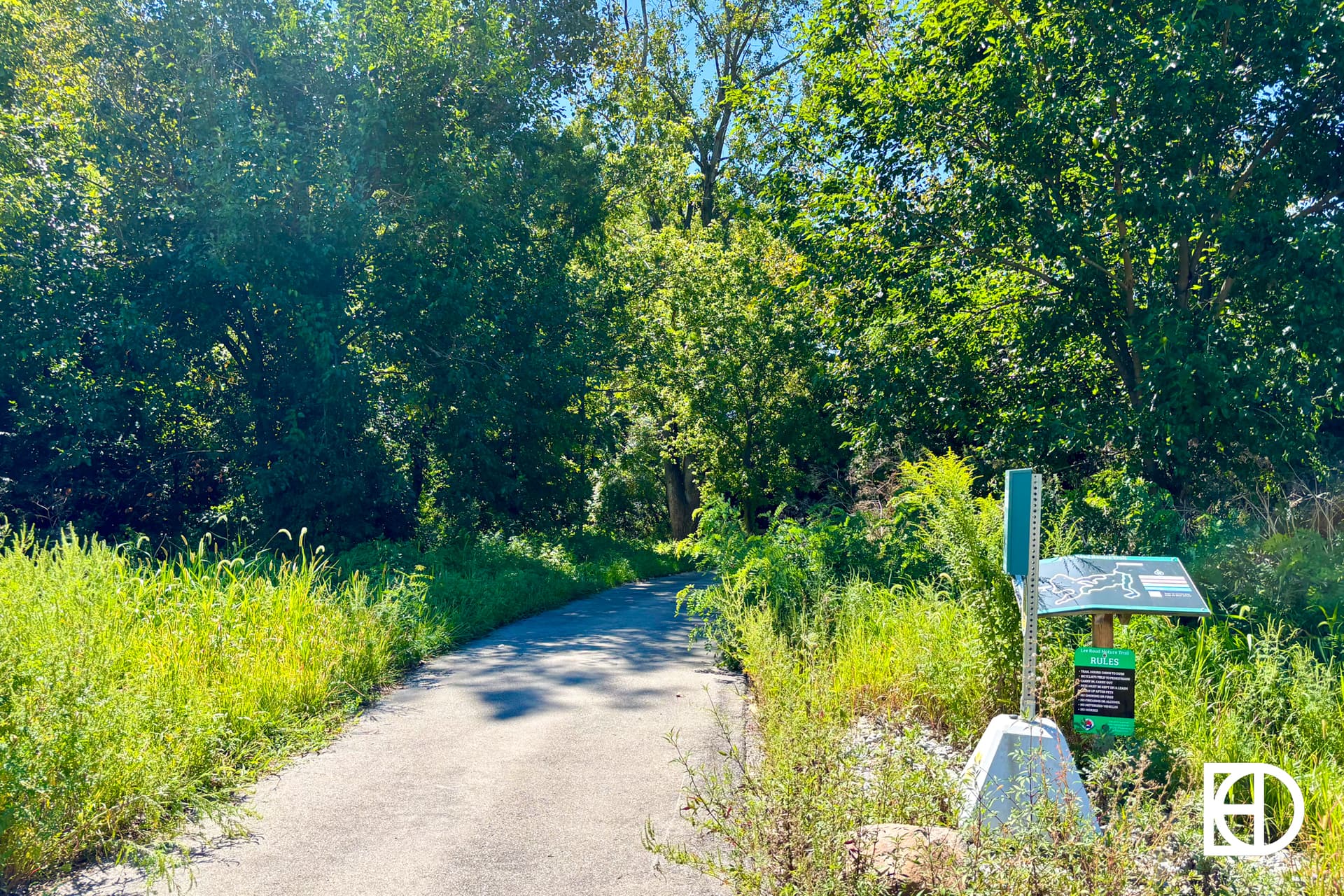 A paved path curves through lush green trees and tall grass on a sunny day; a sign with maps and information stands to the right of the path.