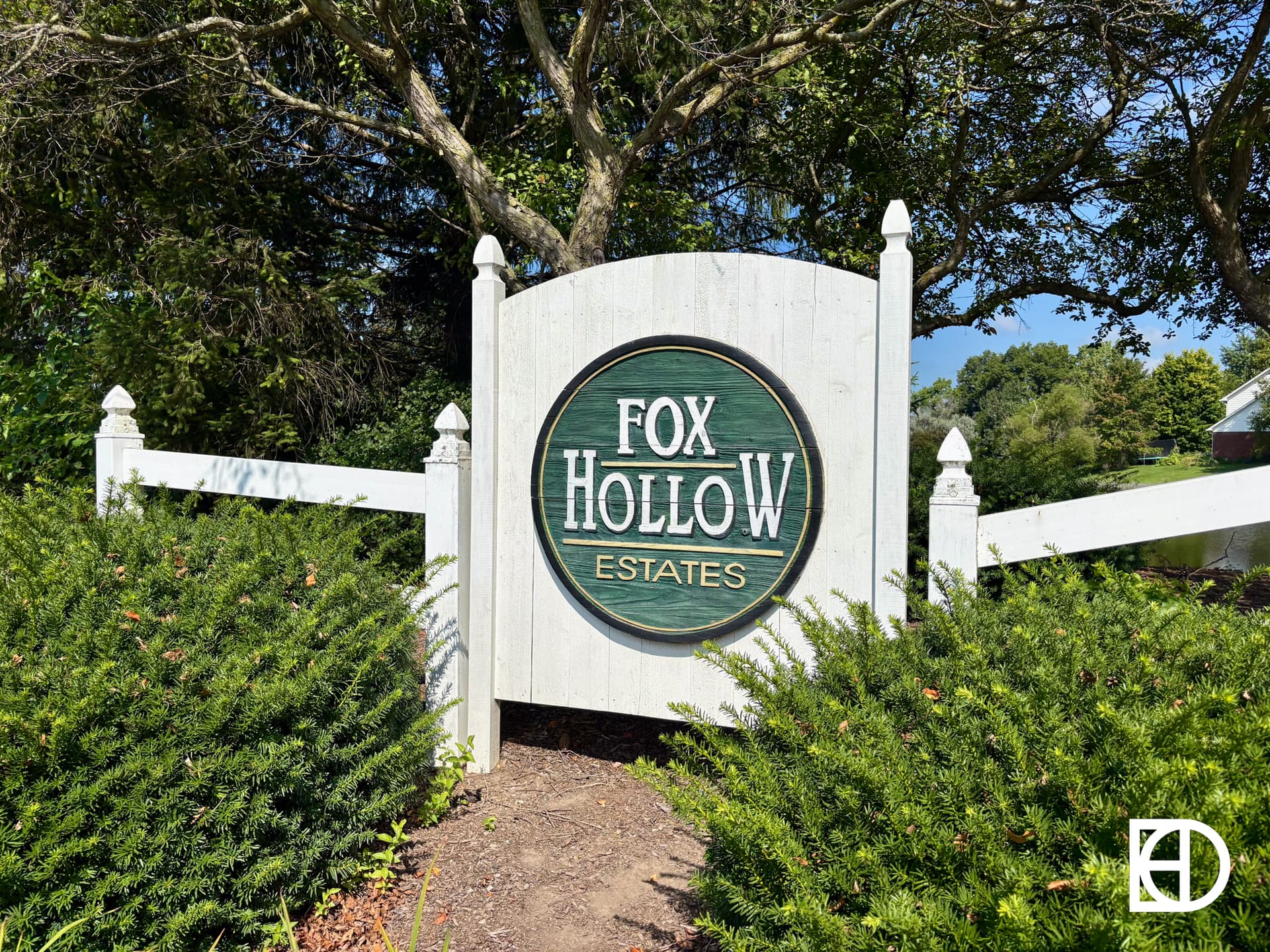 A white wooden sign with a green circular center reads Fox Hollow Estates, surrounded by bushes and trees on a sunny day. The KD logo is in the bottom right corner.