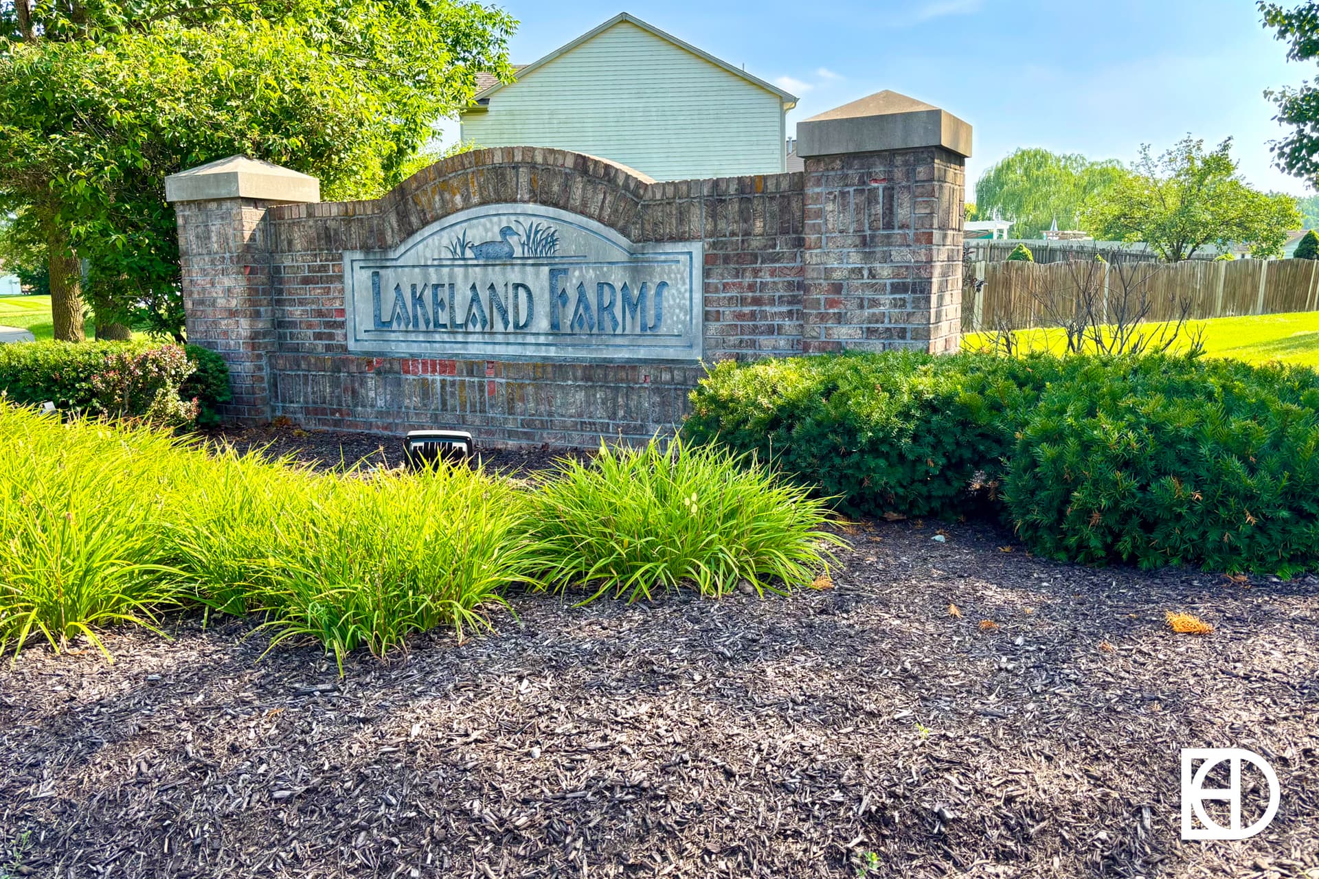 A brick entrance sign for Lakeland Farms is surrounded by green bushes and grass, with trees and houses in the background on a sunny day. The sign features decorative stonework and landscaping.