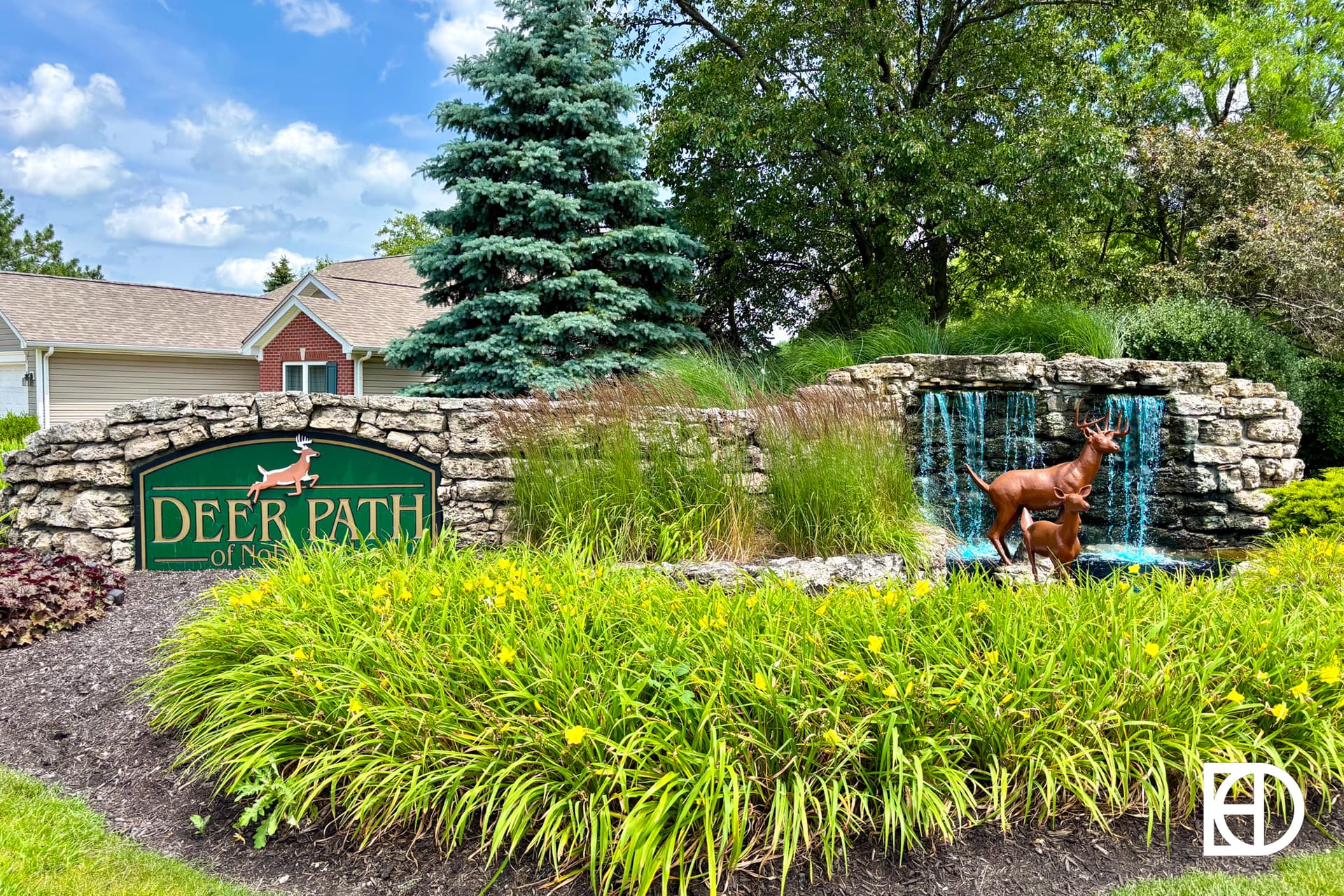 A landscaped entrance sign for Deer Path of North Aurora features a stone wall, a statue of two deer, a small waterfall, and lush greenery with yellow flowers in front of residential homes.