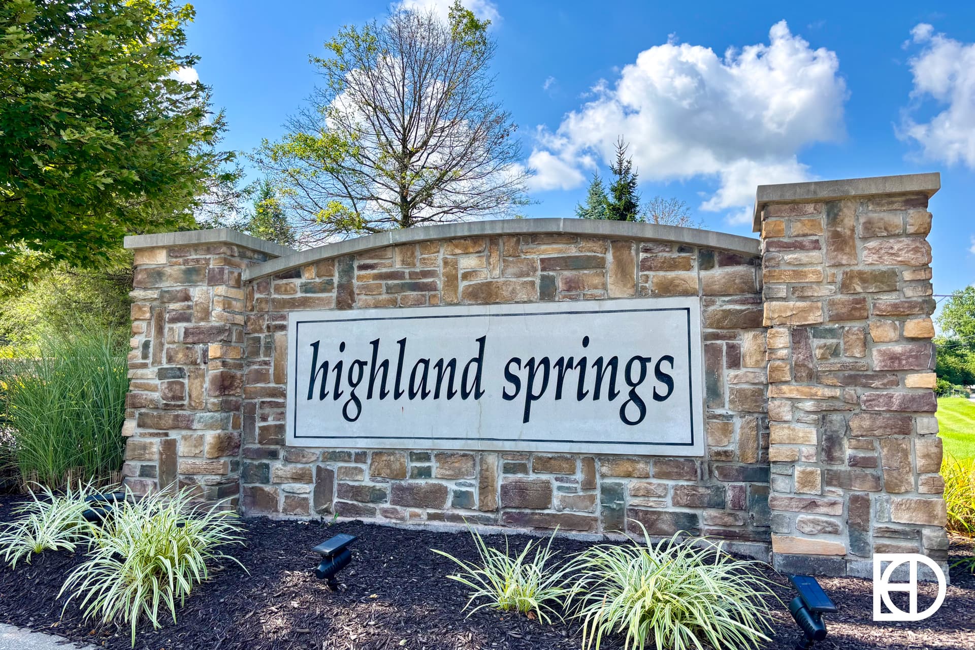 A stone entrance sign with the words highland springs stands in landscaped greenery under a sunny, partly cloudy sky.