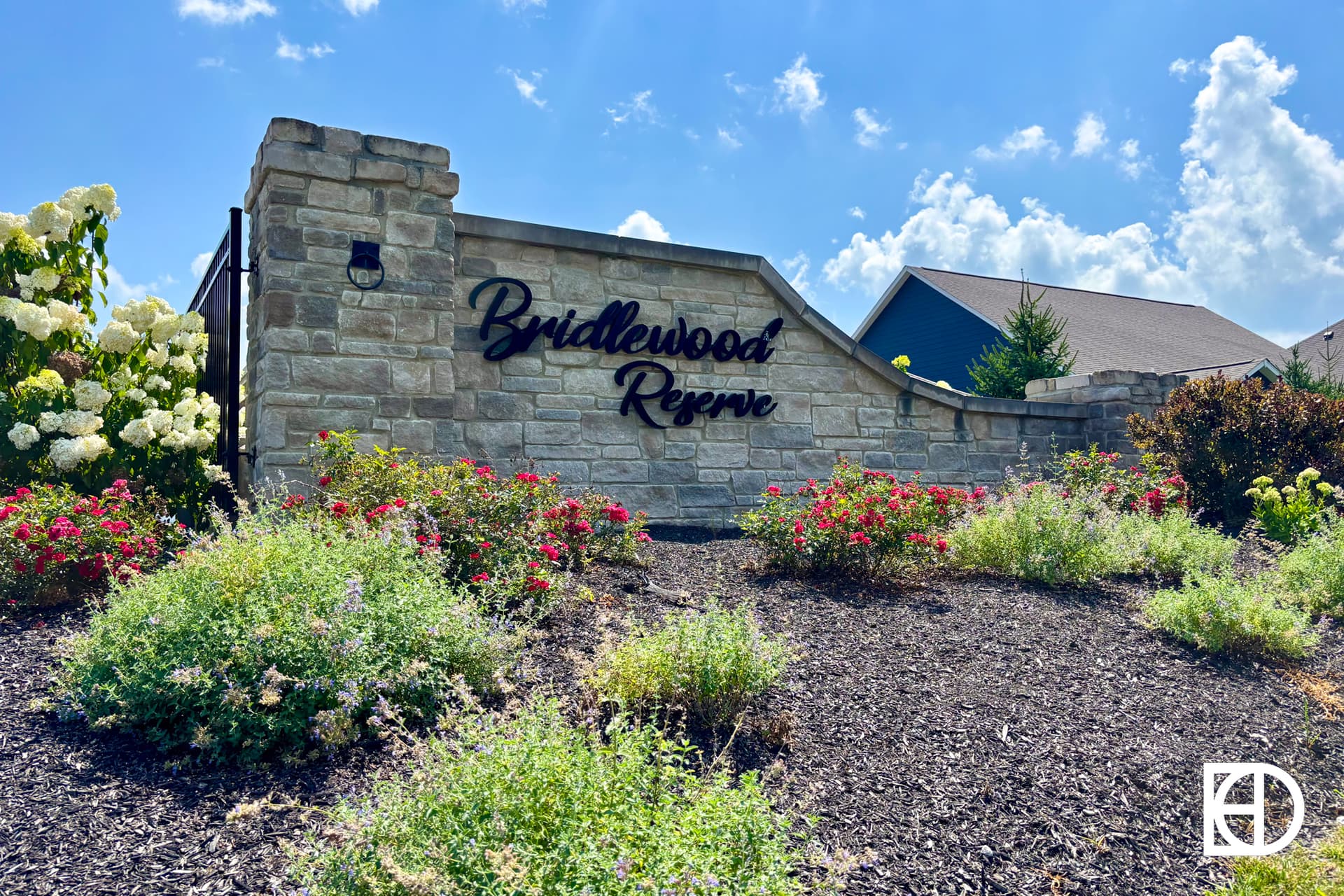 A stone entrance sign reads Bridlewood Reserve, surrounded by blooming flowers, shrubs, and mulch, with houses and a partly cloudy blue sky in the background.