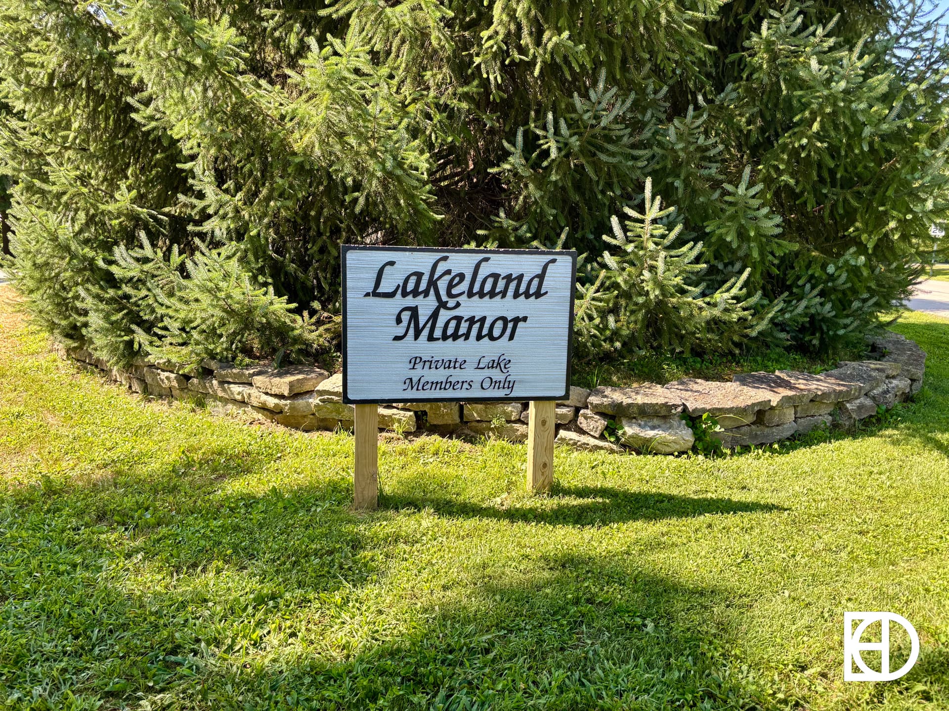 A wooden sign on grass reads Lakeland Manor, Private Lake, Members Only, with trees and a stone-edged garden in the background.