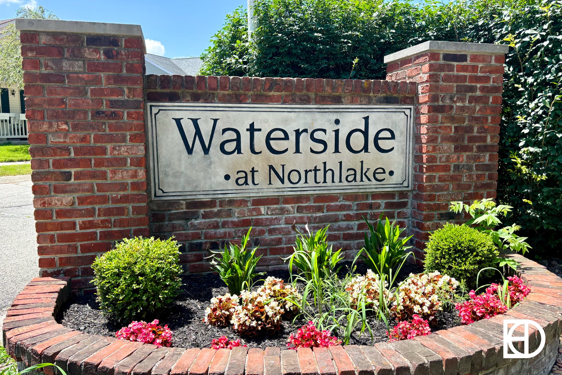 A brick sign with engraved stone reads Waterside at Northlake, surrounded by green bushes and blooming flowers, with a house and blue sky in the background.